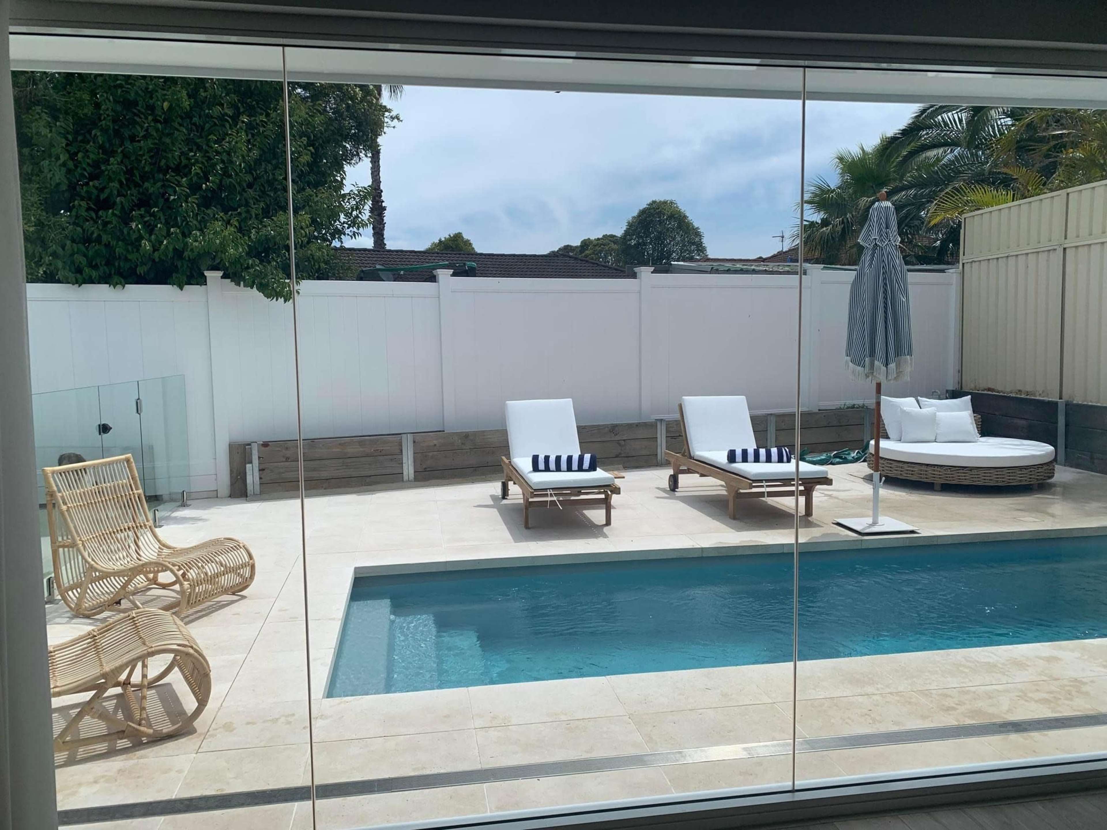 An outdoor area with a swimming pool, two lounge chairs, a round daybed, and a sun umbrella, all surrounded by a white wall and greenery.