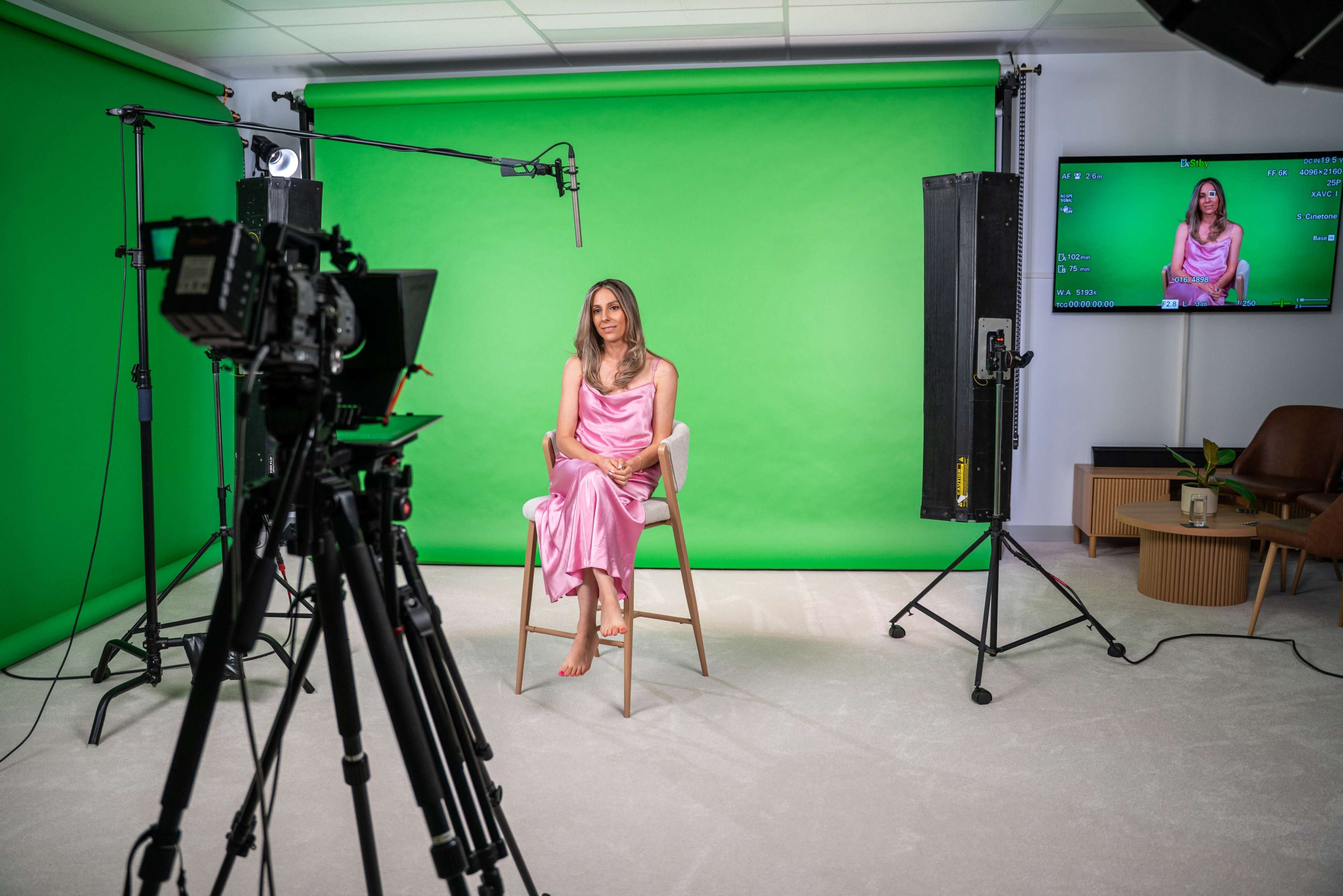 A woman in a pink dress sits on a stool in front of a green screen, with cameras and lighting equipment set up around her.