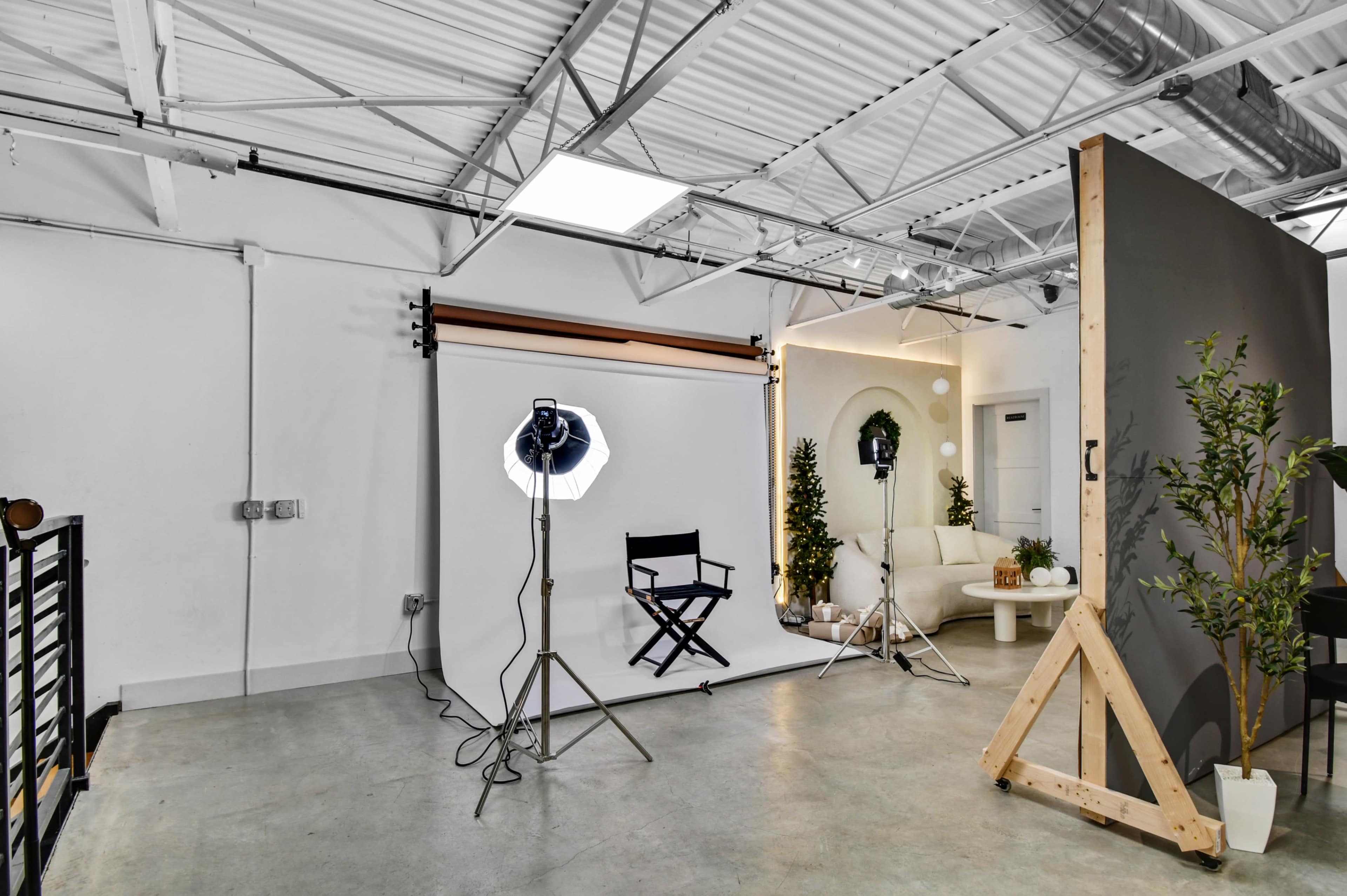 The image shows a photography studio with a white backdrop, lighting equipment, a director's chair, and a sofa against a minimalist interior.