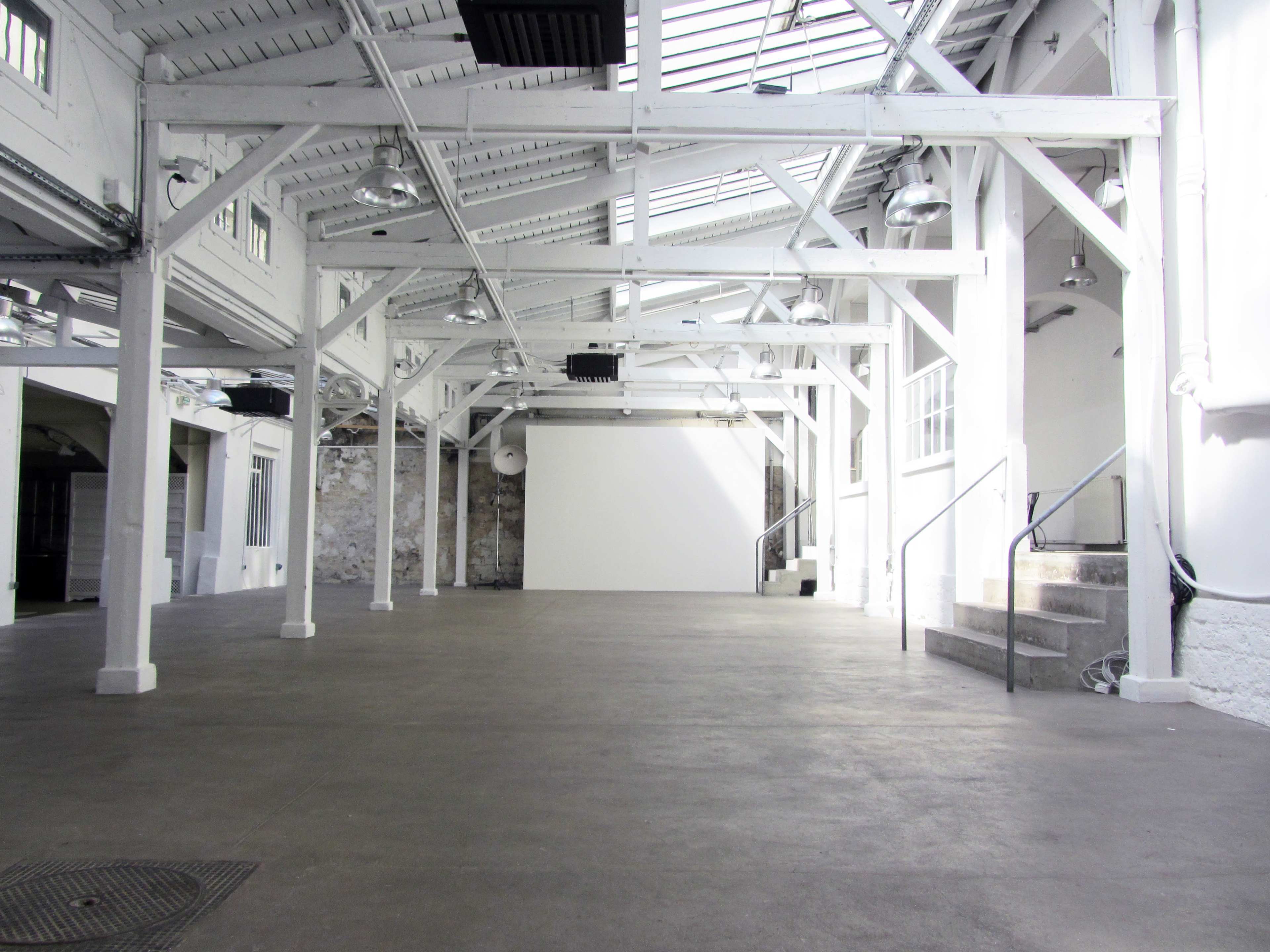 A spacious, empty industrial hall with white walls, wooden beams, and a large blank wall at the far end.