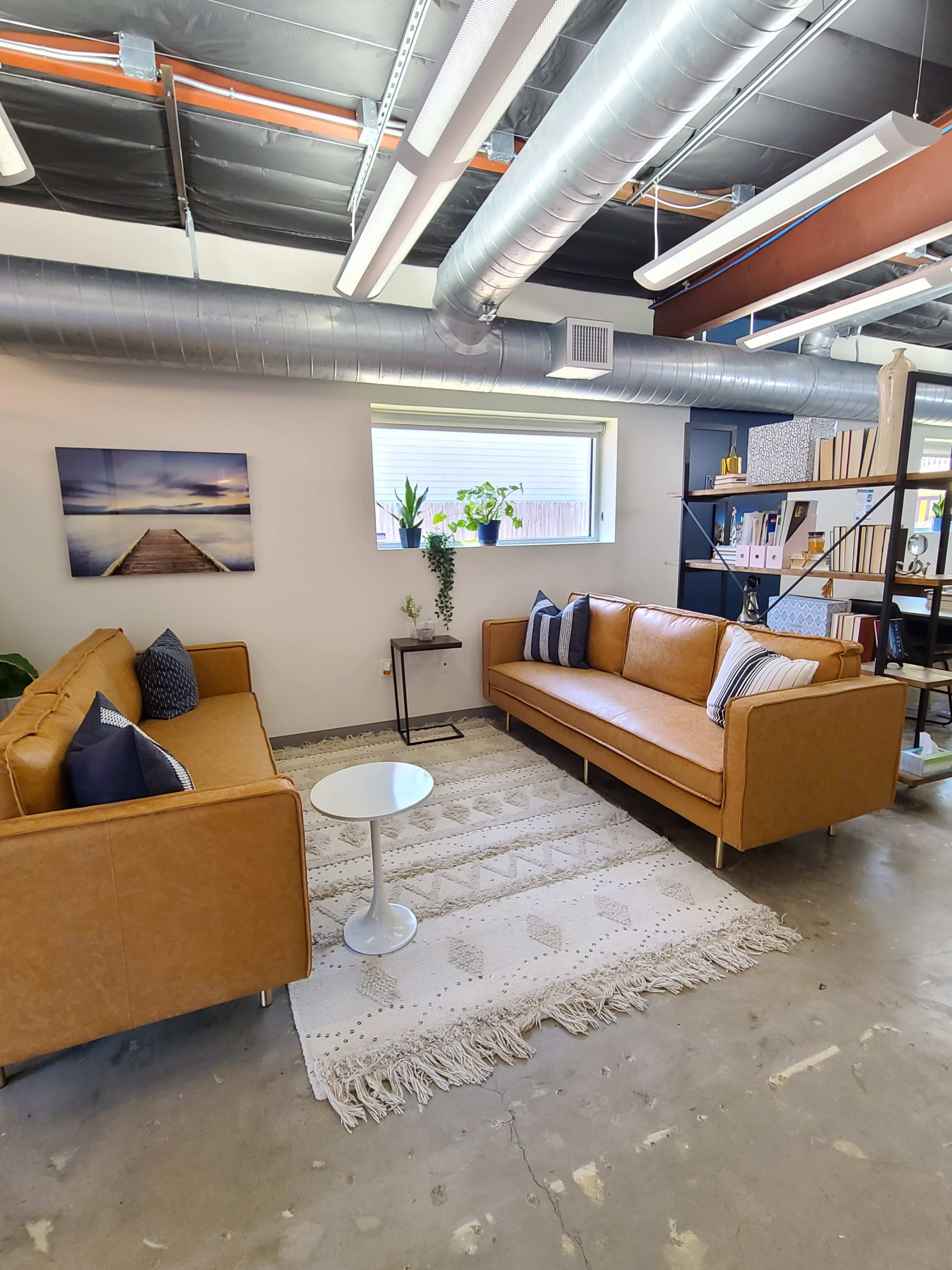 A modern office space featuring two brown leather sofas, a white side table, and a large rug, with a window providing natural light.