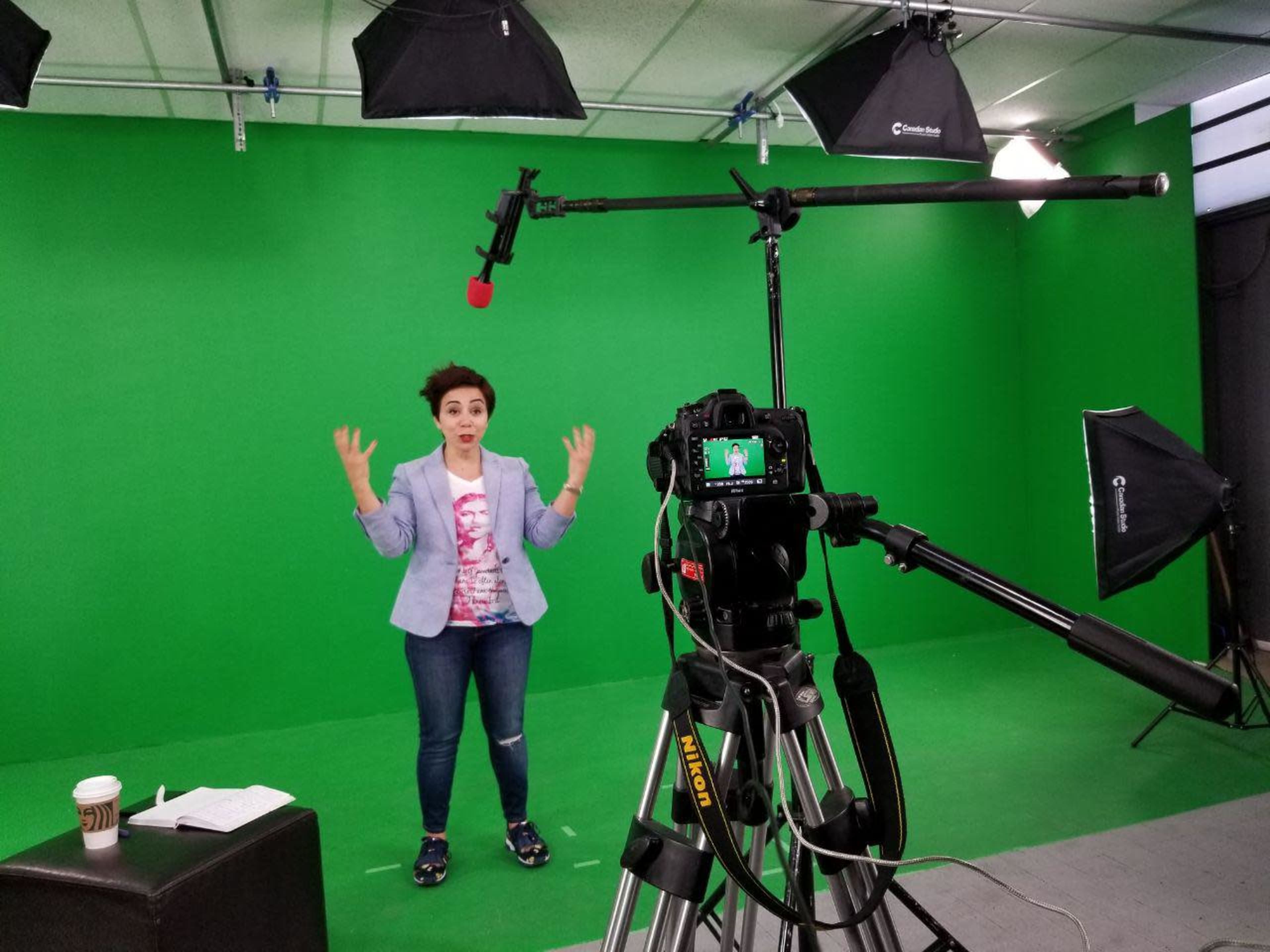 A person stands in front of a green screen, gesturing while facing a camera mounted on a tripod.