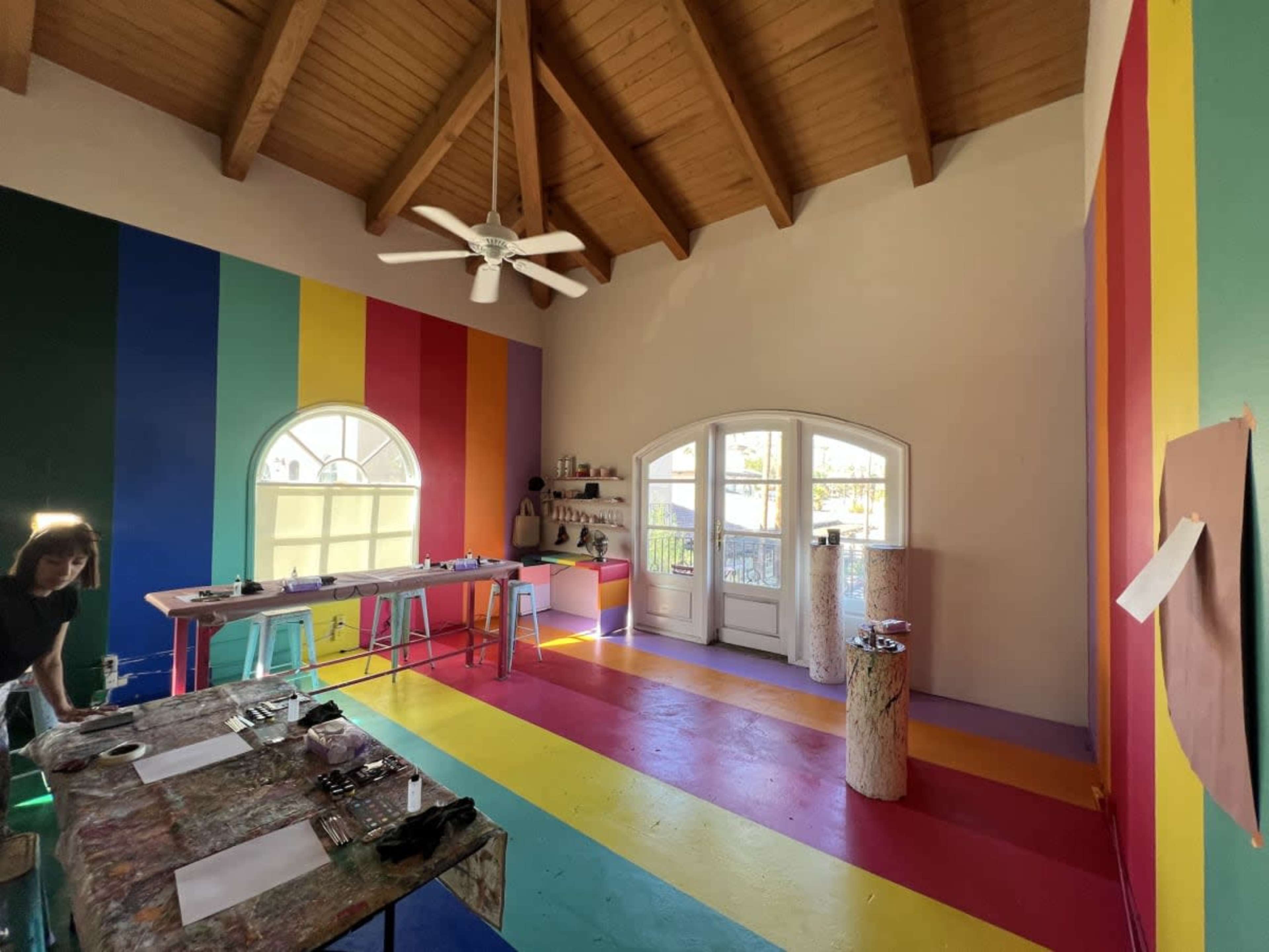 The image shows a brightly colored studio space with rainbow stripes on the floor and walls, featuring a central round window and several art supplies on a table.
