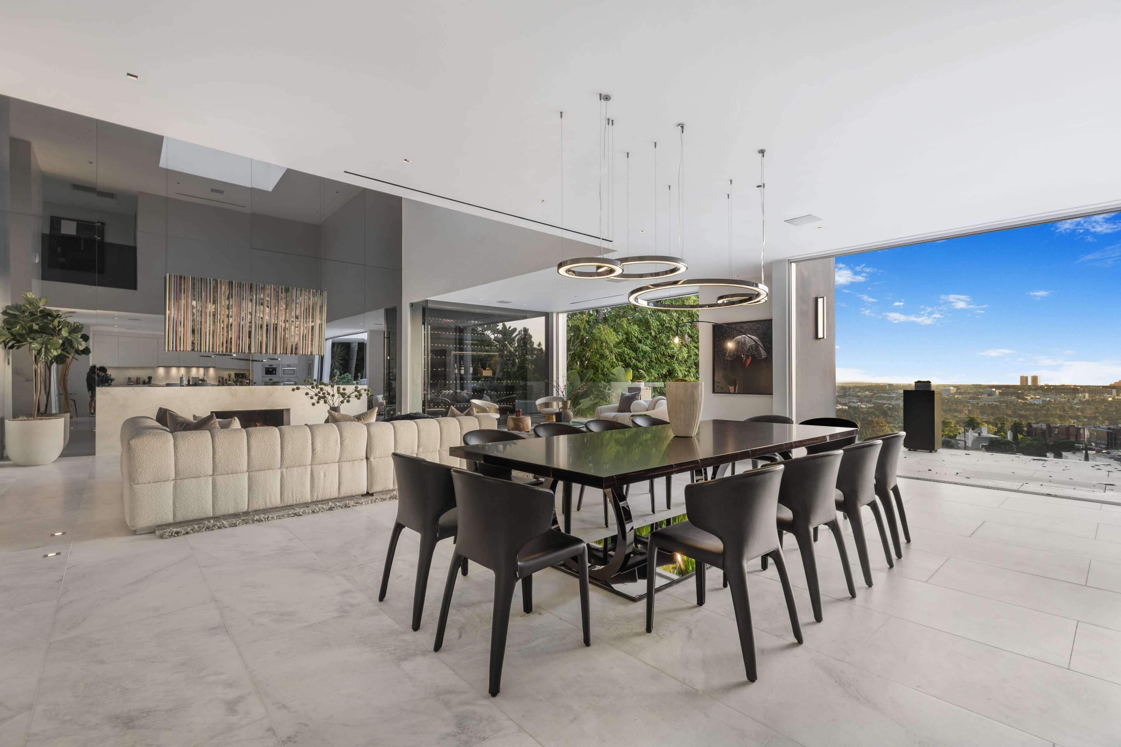 The image shows a modern, open-concept dining area with a large table surrounded by black chairs, large windows offering a view of the outdoors, and sleek, contemporary furnishings.