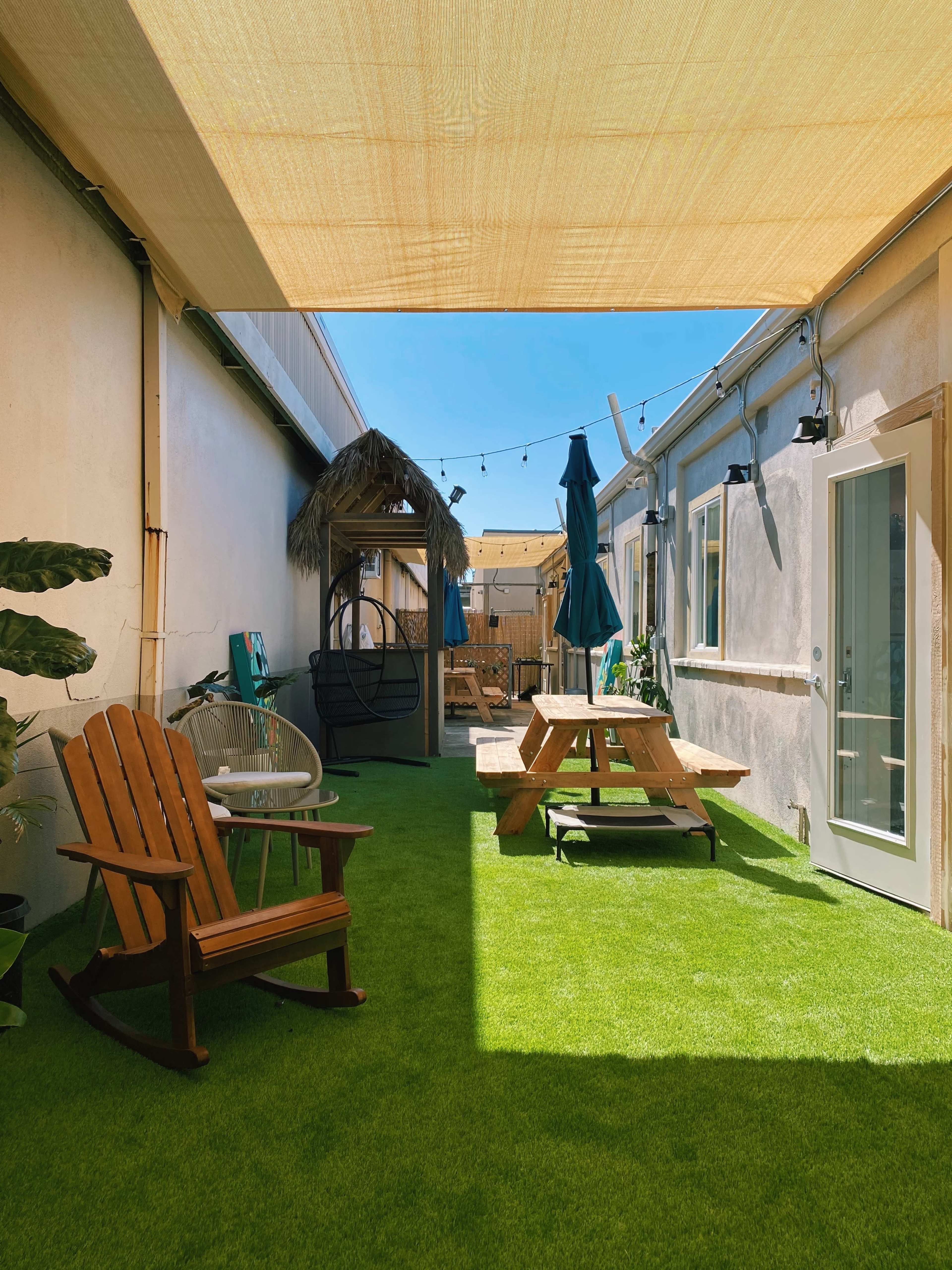 A well-lit outdoor courtyard with artificial grass, featuring a rocking chair, a picnic table, a shaded area with a thatched roof, and string lights.
