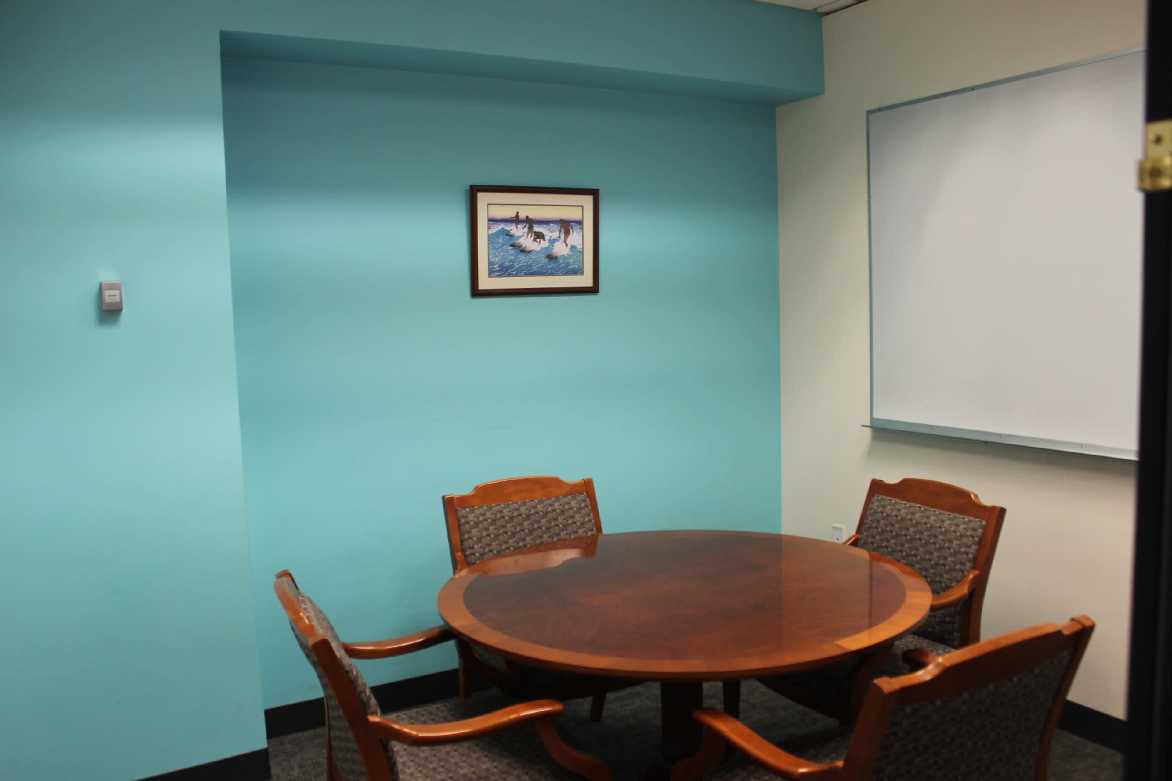 A round wooden table surrounded by four wooden chairs is positioned in a turquoise-colored meeting room with a framed picture on the wall and a whiteboard.