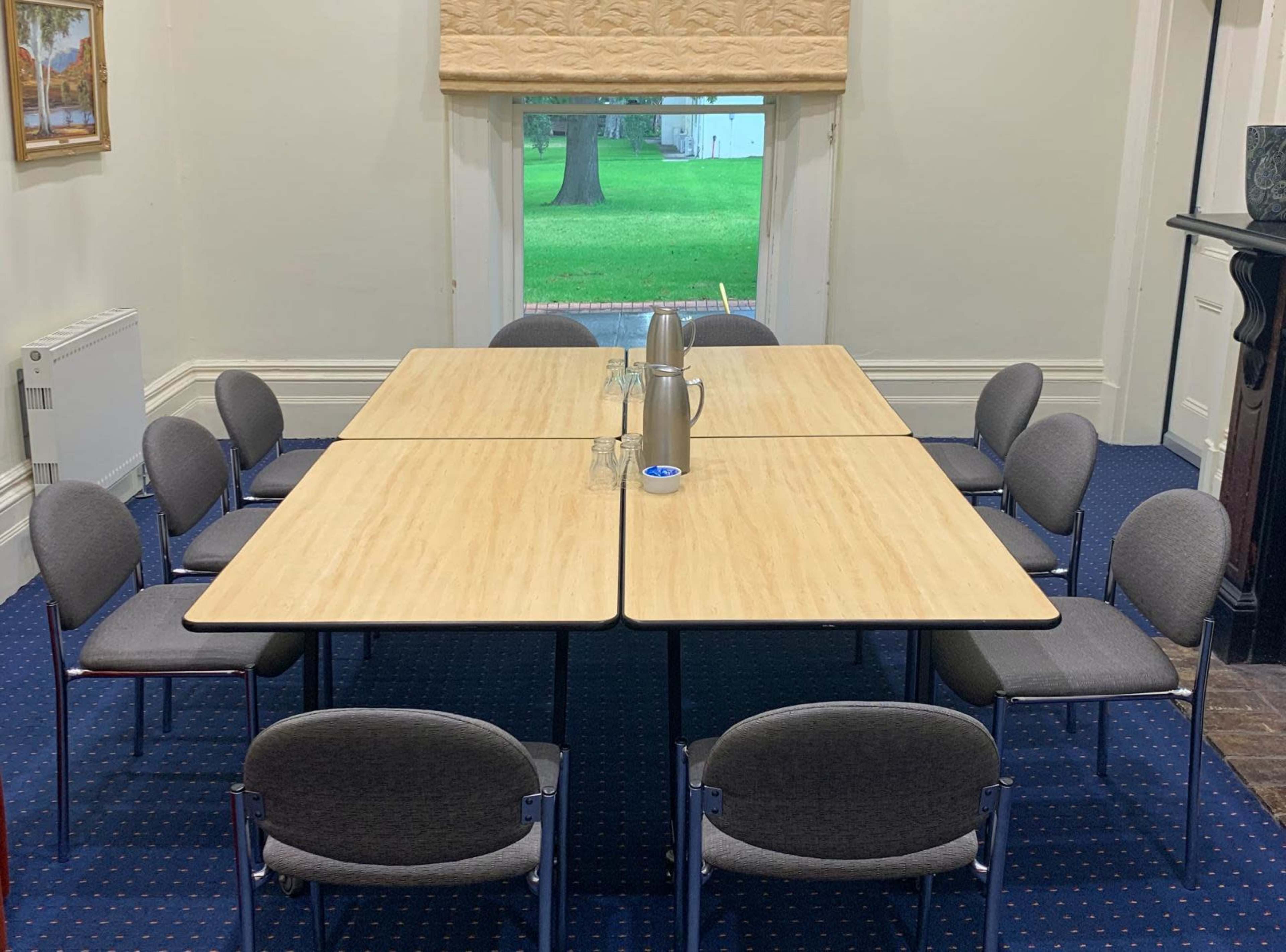 A rectangular conference table is set in a meeting room with ten gray chairs arranged around it, featuring two water pitchers and glasses placed in the center.