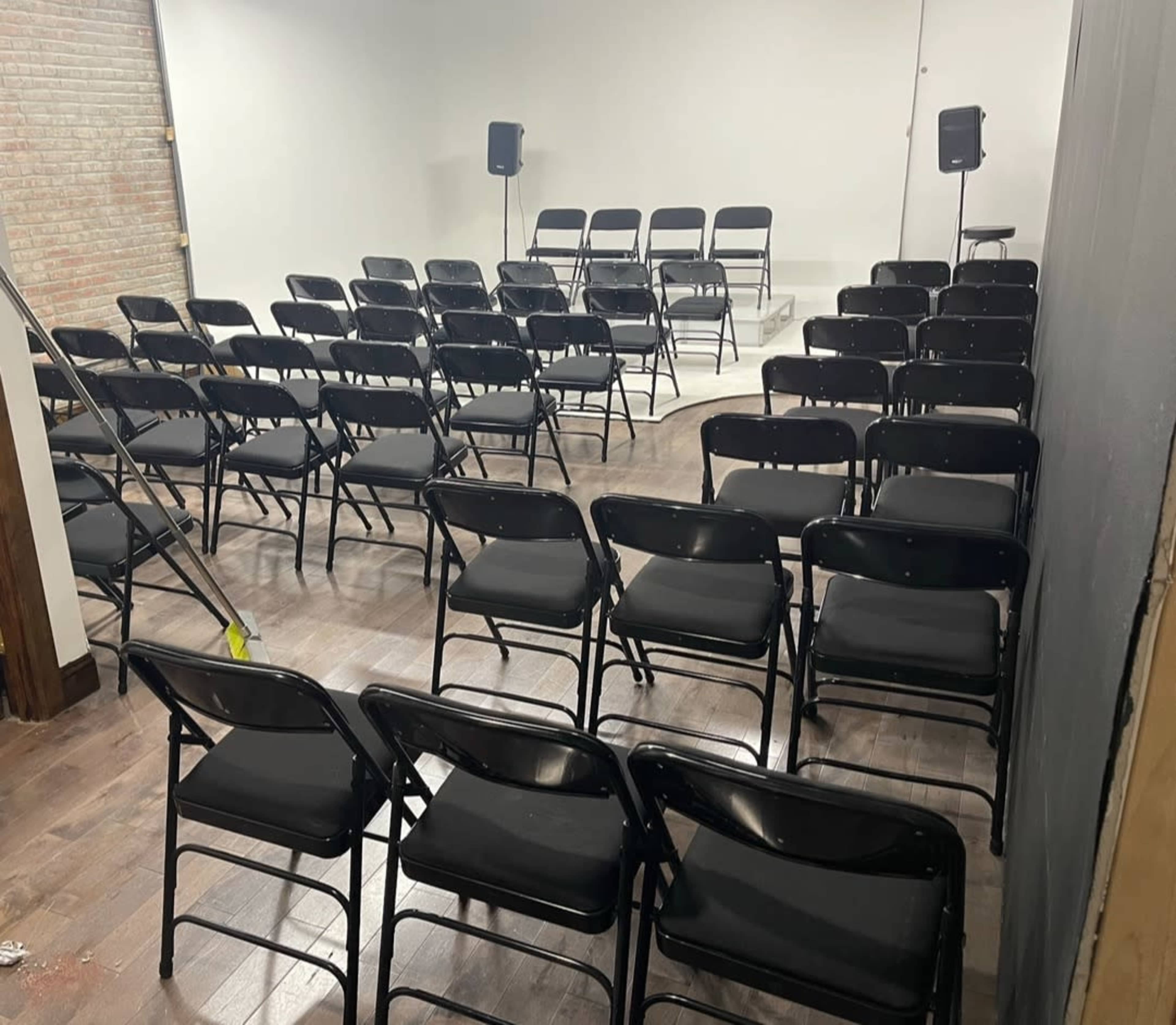 A row of black folding chairs is set up in a partially empty room with a light-colored wall and speakers positioned at the front.