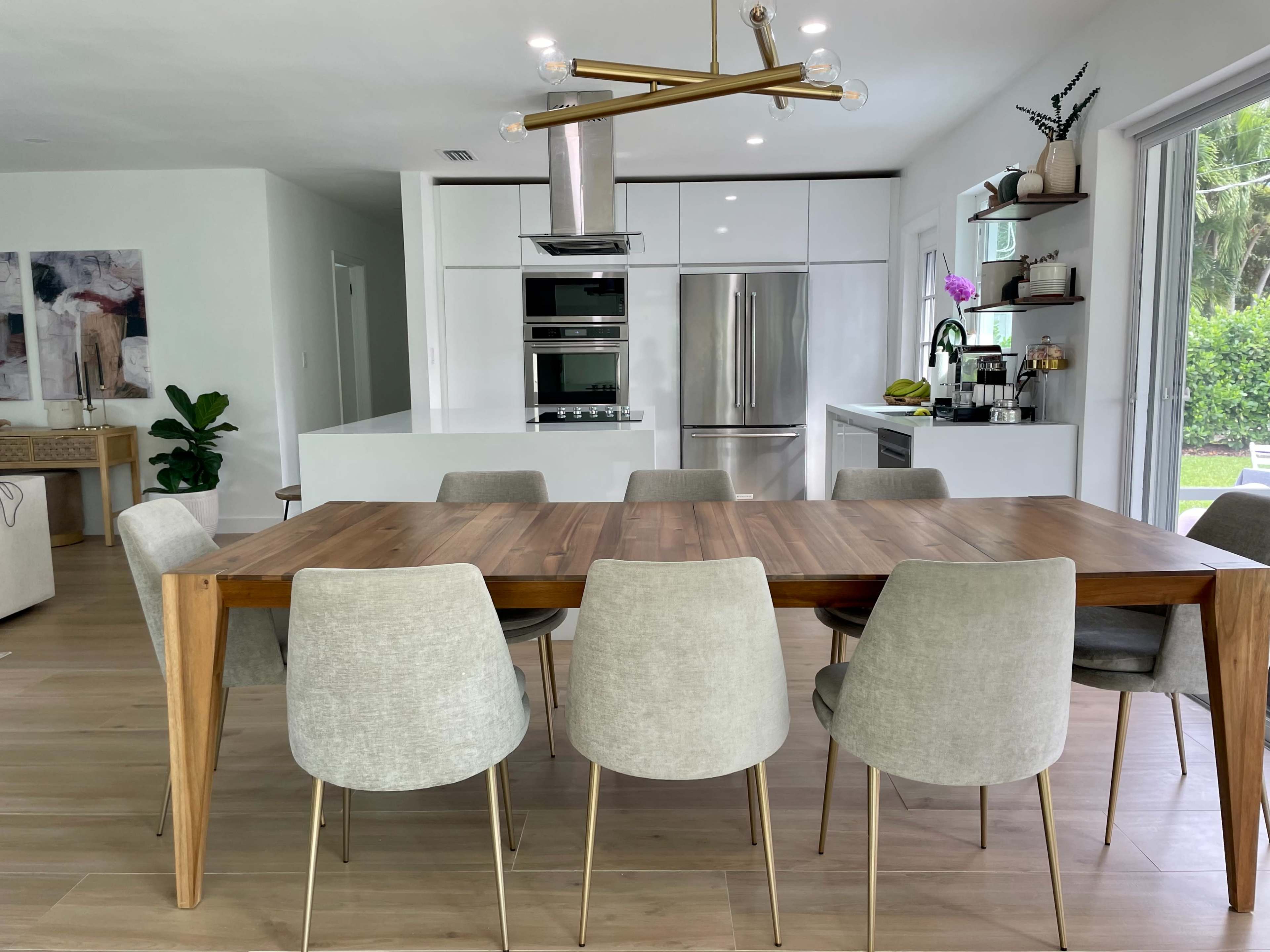 The image shows a modern kitchen and dining area featuring a wooden dining table surrounded by gray upholstered chairs, with sleek cabinetry and stainless steel appliances in the background.