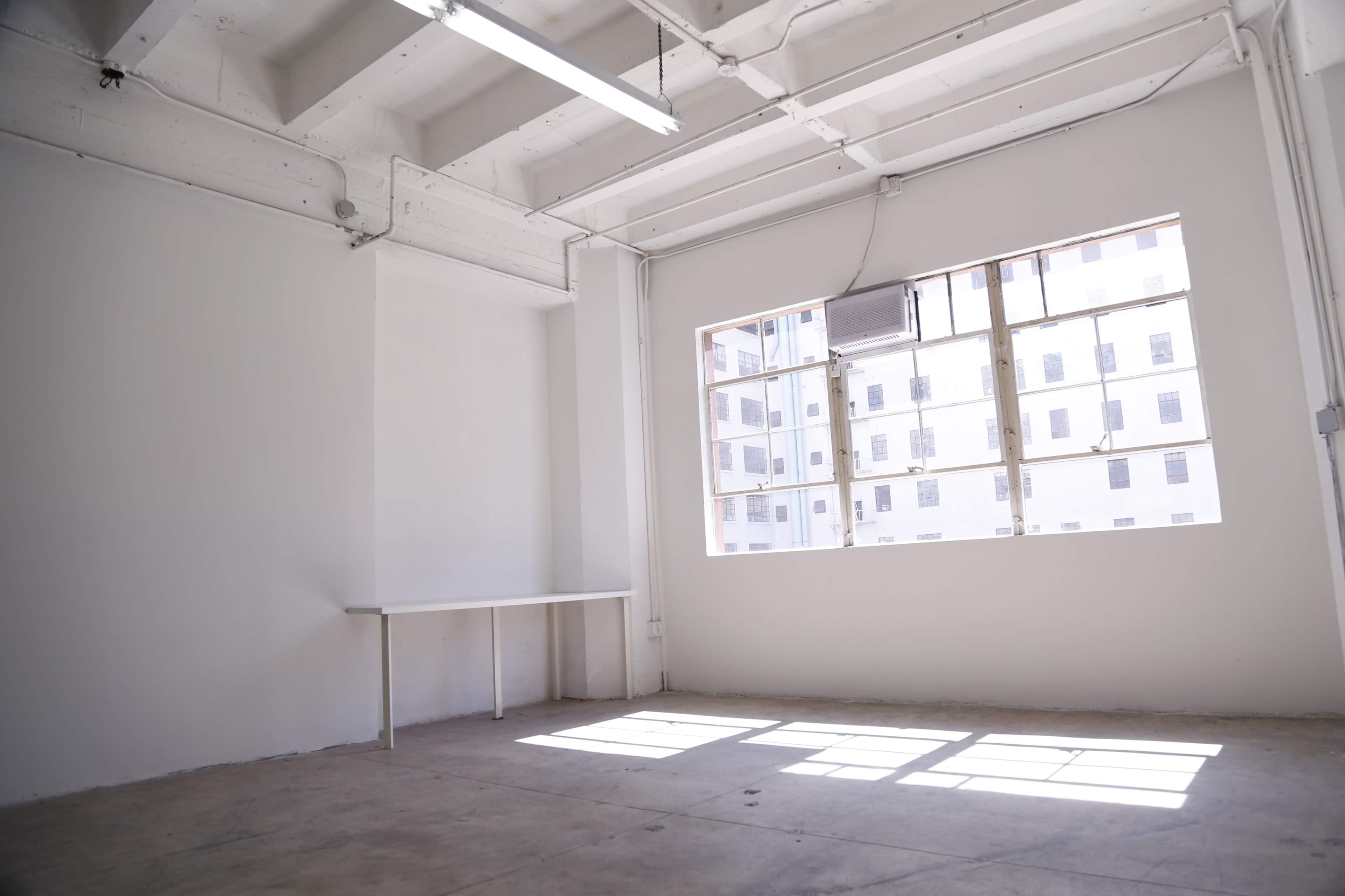 The image shows an empty room with white walls and a large window letting in natural light, featuring a simple table against one wall.