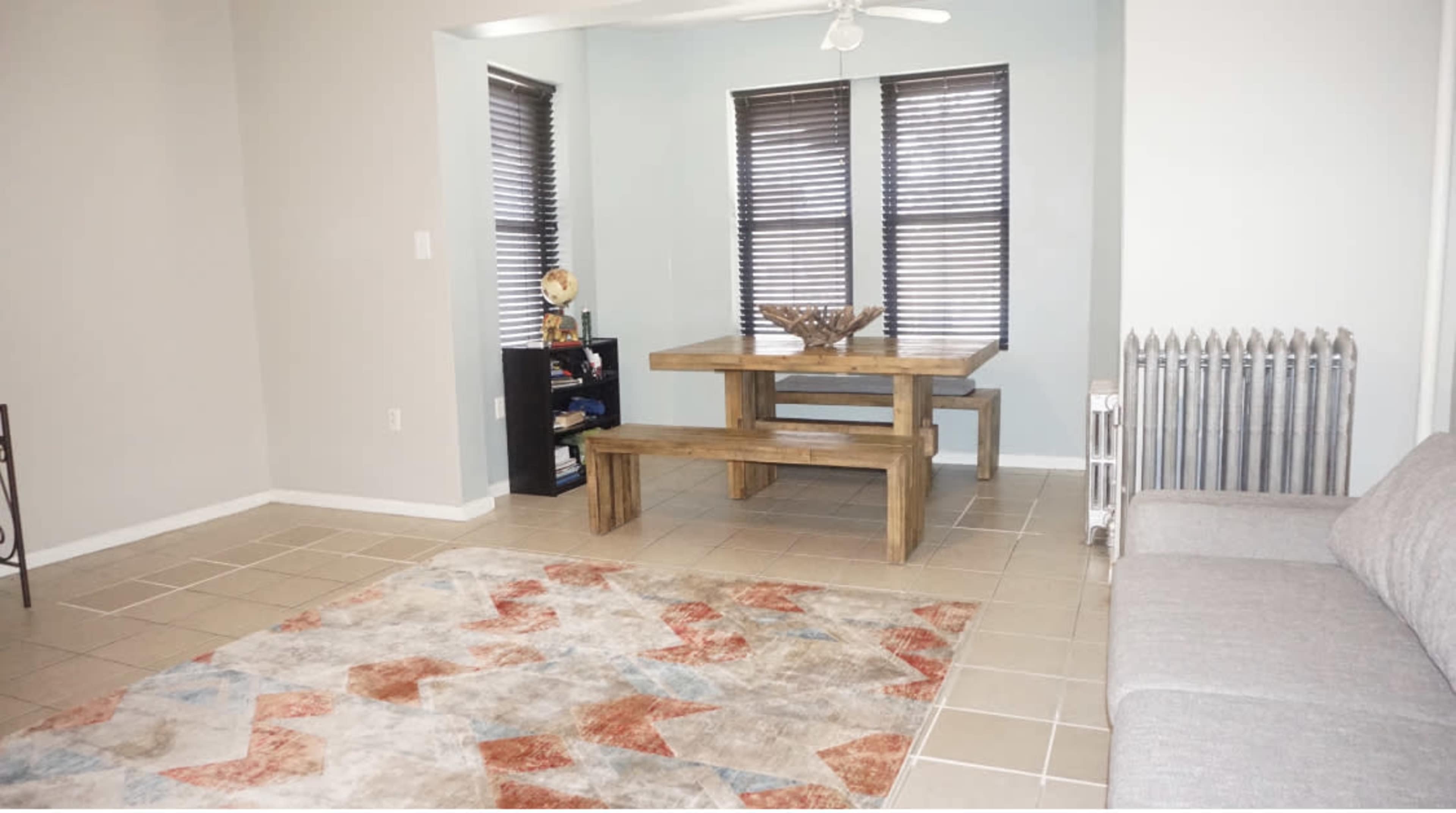 A living area featuring a wooden dining table with a decorative bowl, a gray couch, a patterned rug, and window blinds.
