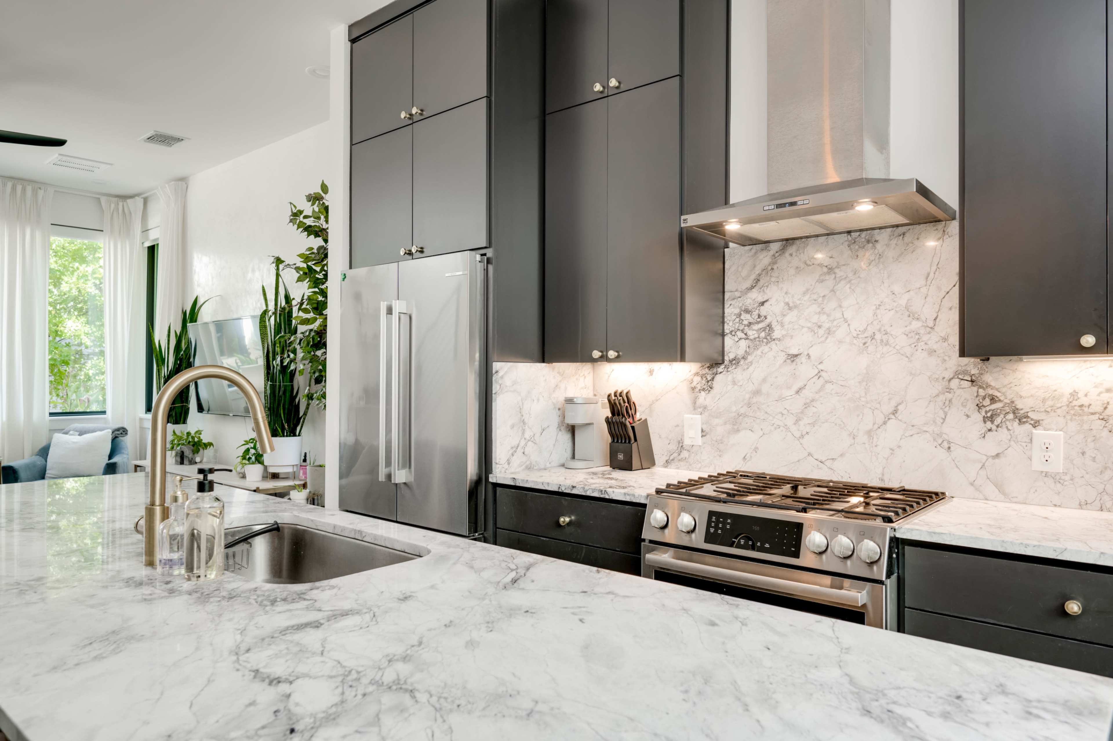 The image shows a modern kitchen featuring dark cabinetry, a marble countertop, a stainless steel gas stove, and a large sink with a sleek faucet.