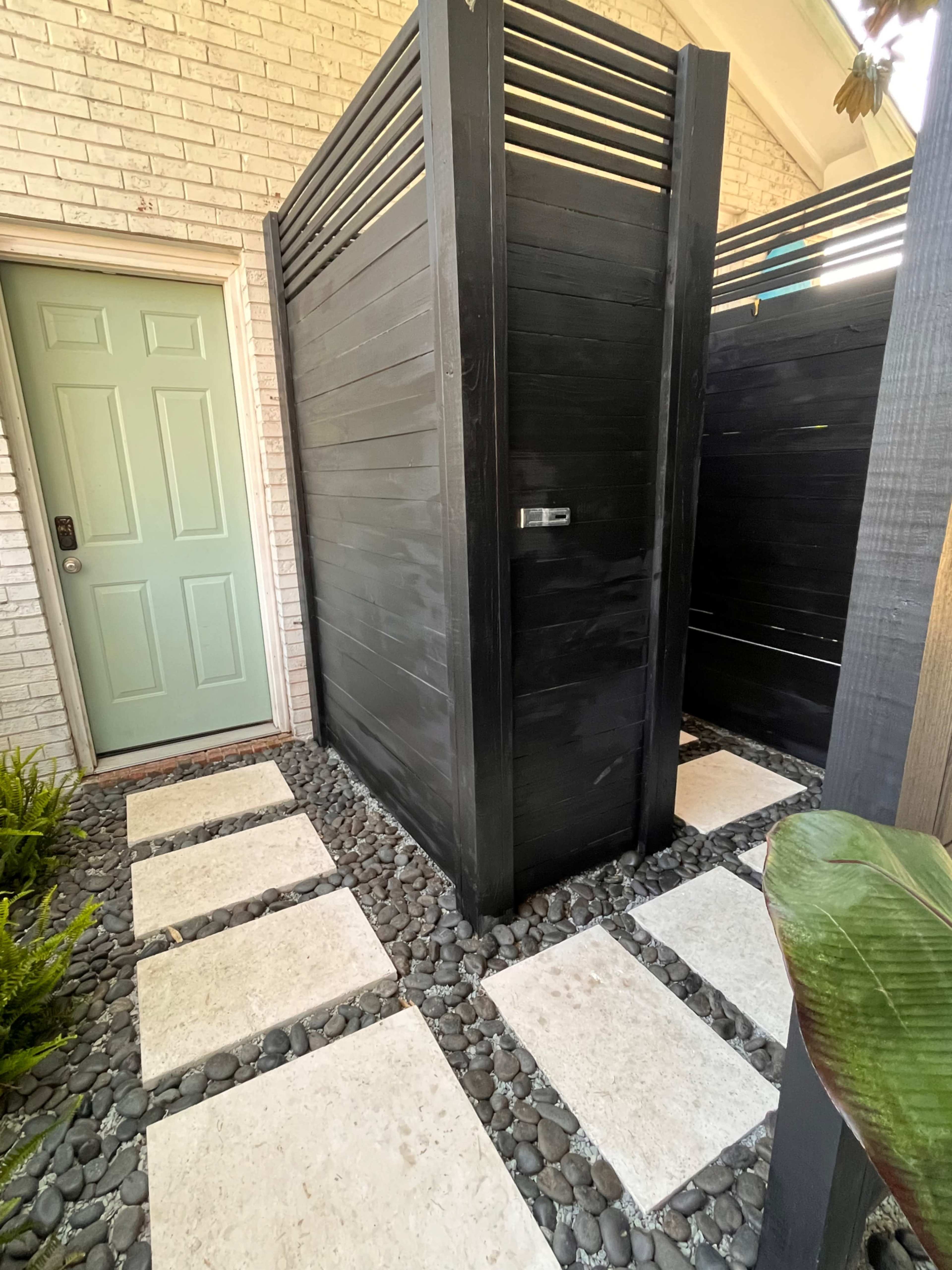 An outdoor shower area with two wooden privacy enclosures, a green door, and a pathway of large stone slabs bordered by small pebbles.