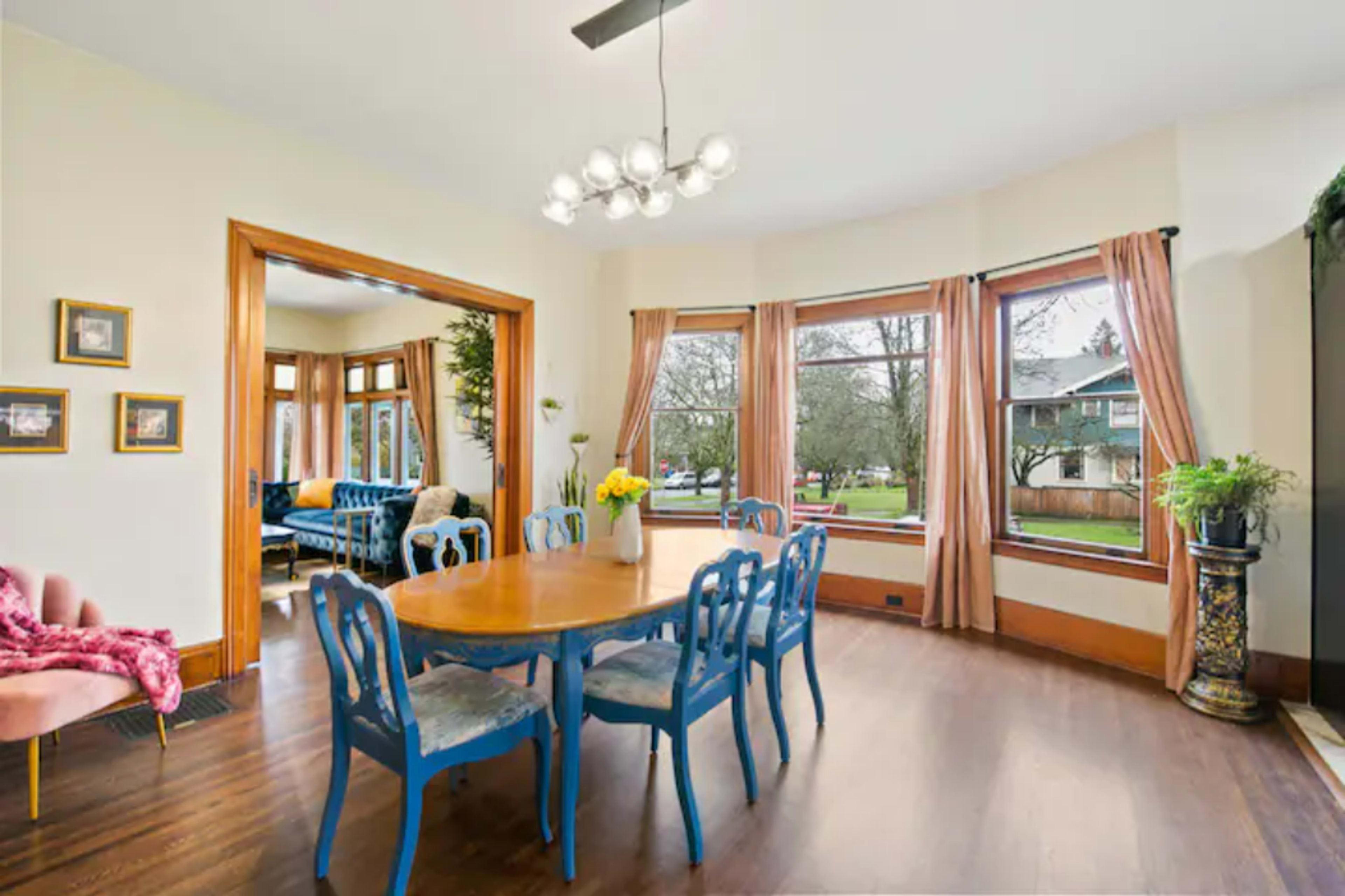 A dining room features a round wooden table surrounded by blue chairs, with large windows offering views of a garden.