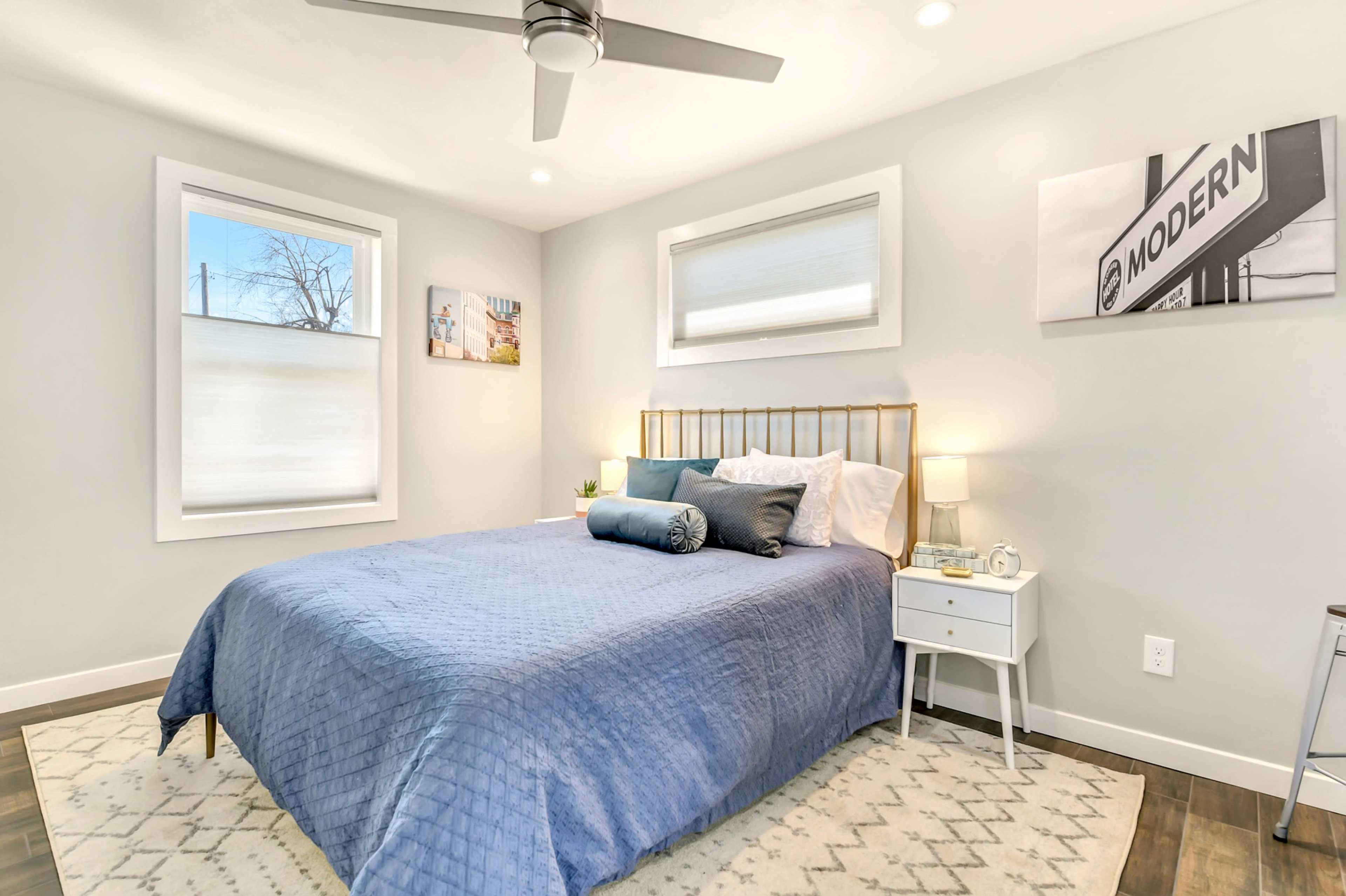 A neatly arranged bedroom featuring a bed with a blue quilt, side tables, and large windows allowing natural light.