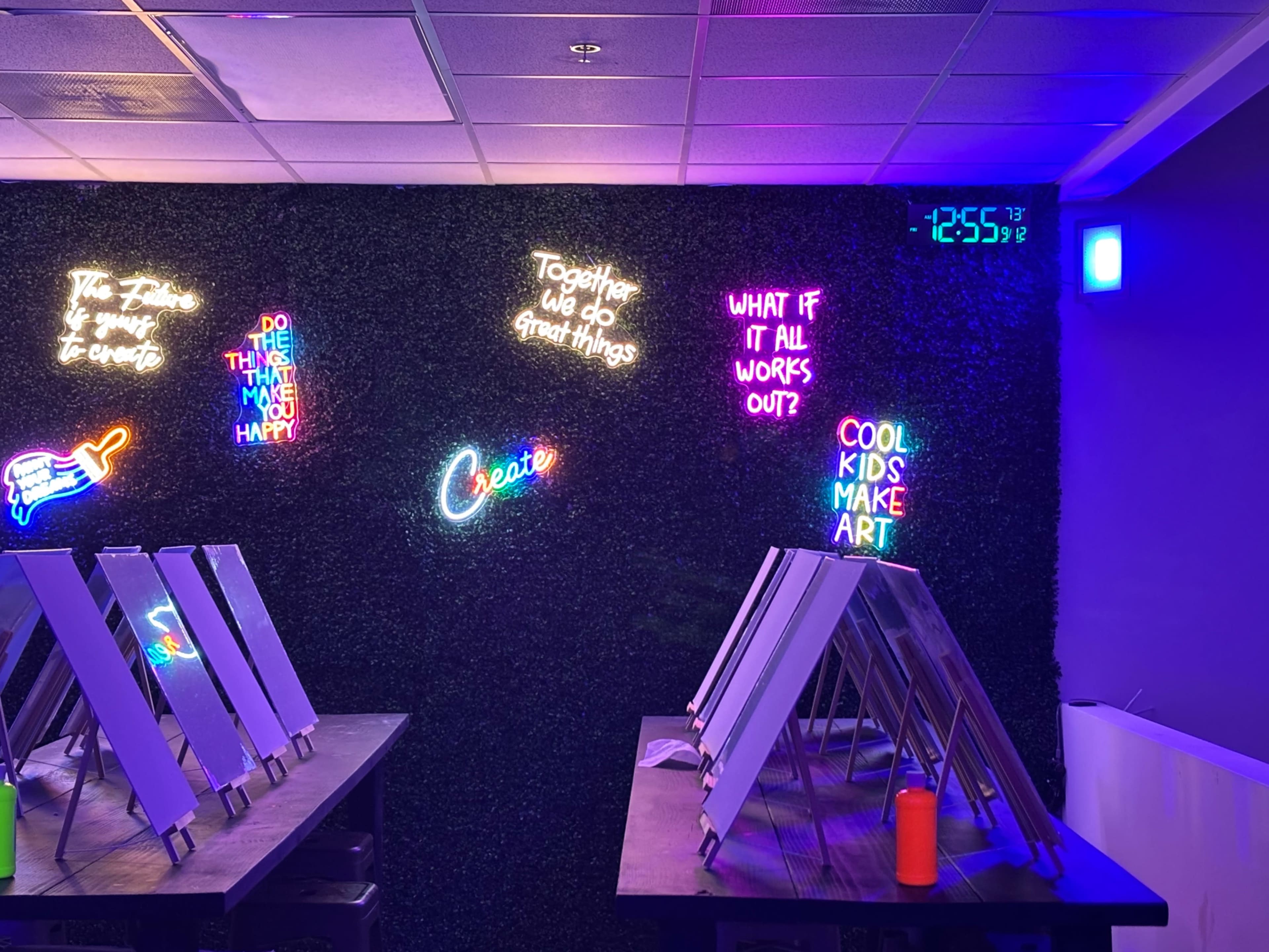 The image shows a wall adorned with colorful neon signs and a digital clock, while tables with easels are positioned in the foreground.