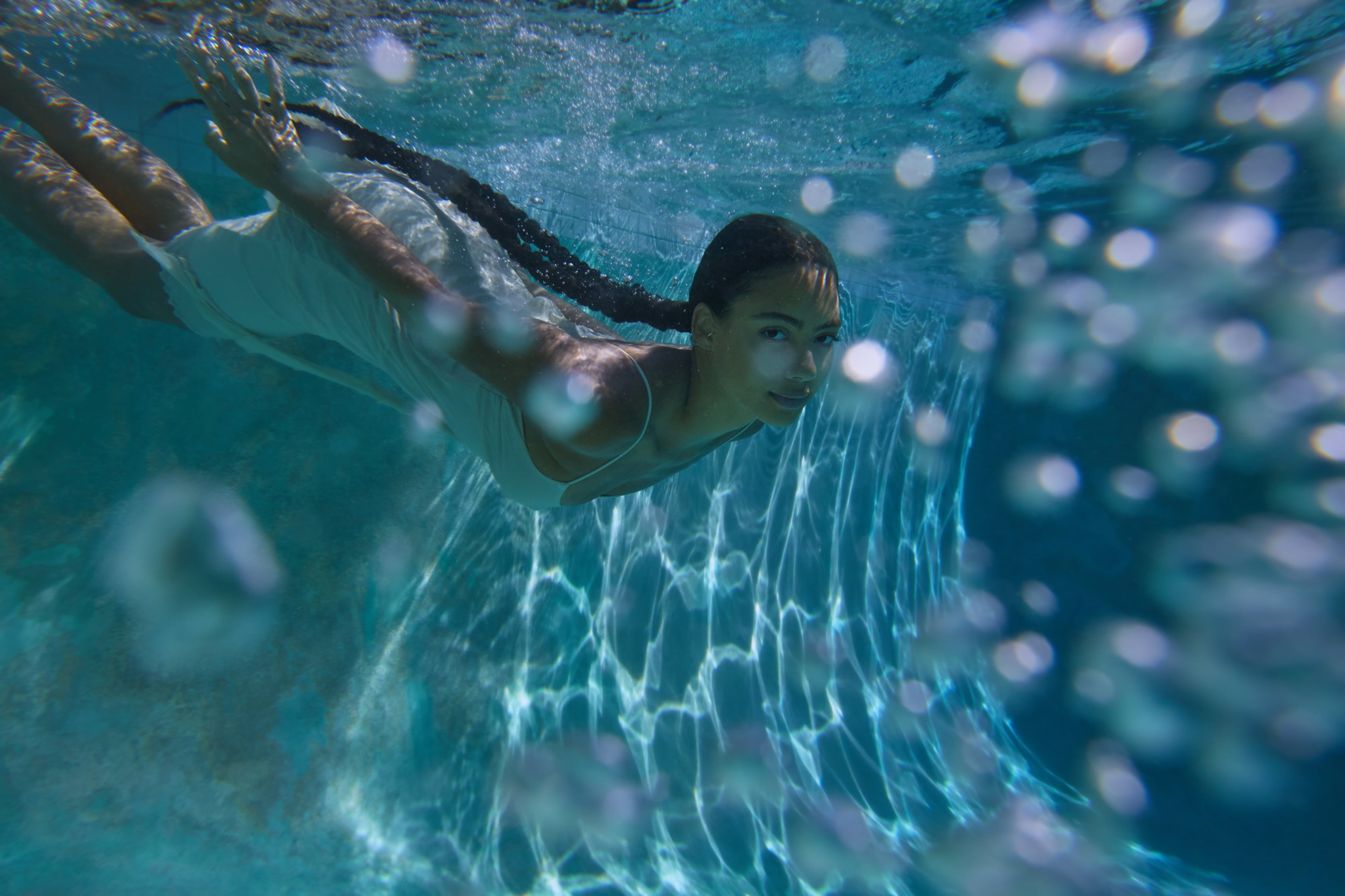 A person is swimming underwater in a pool, with bubbles surrounding them and light reflections on the water's surface.