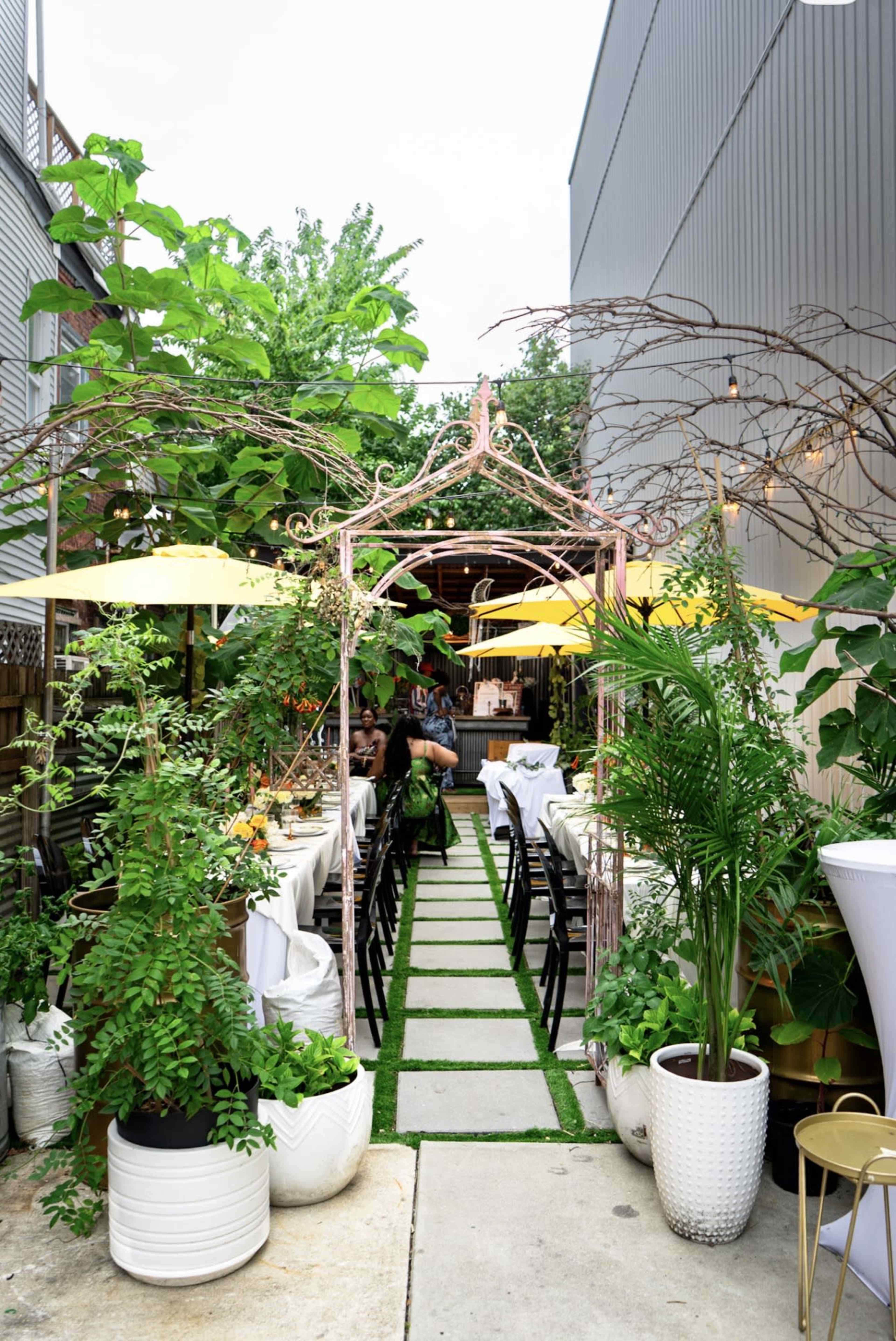 The image shows an outdoor dining area lined with greenery, white tables, and yellow umbrellas, leading to a decorative archway.