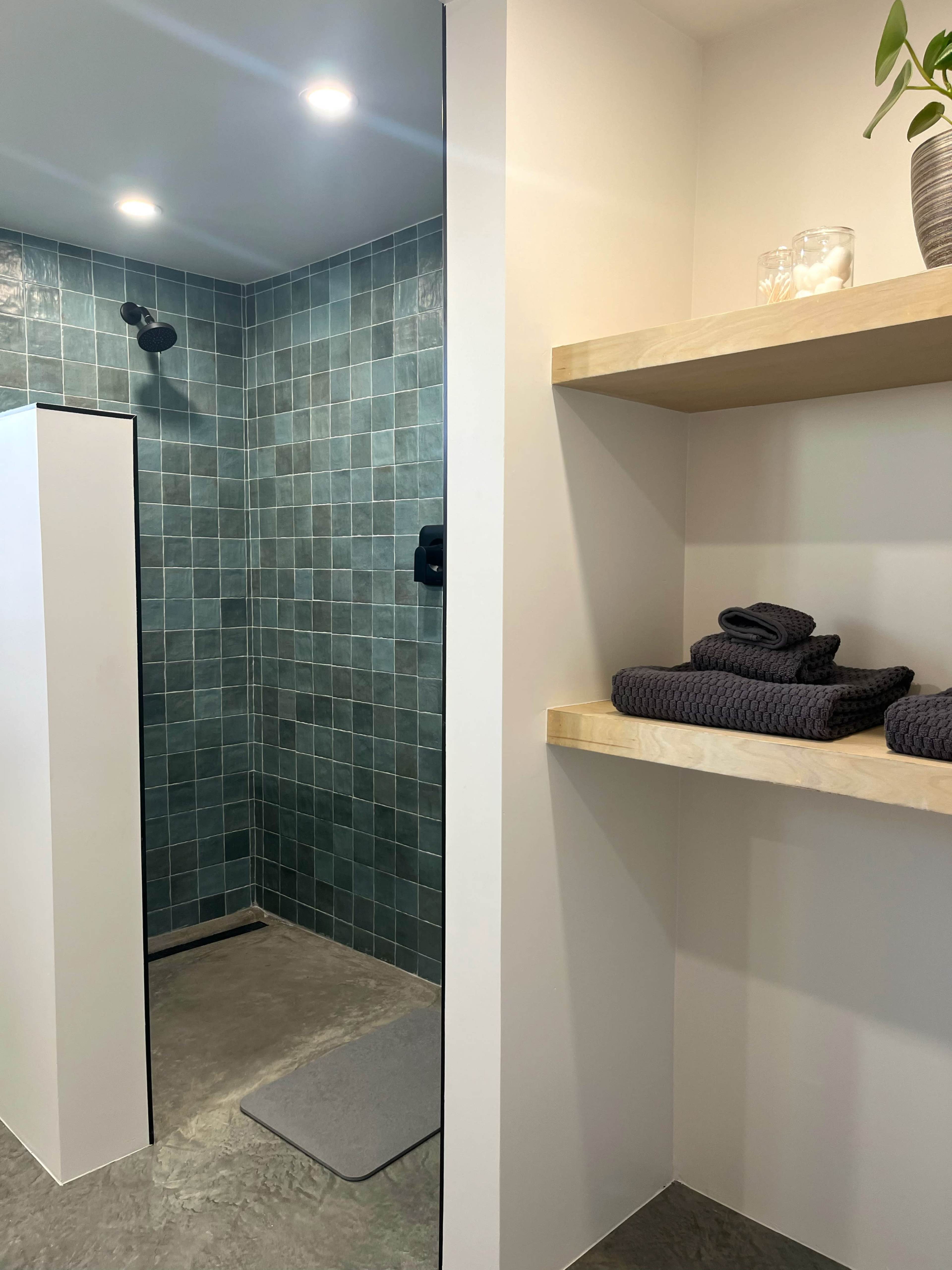 The image shows a modern shower area with teal tiled walls and a glass partition, alongside a wooden shelf displaying neatly folded dark towels.