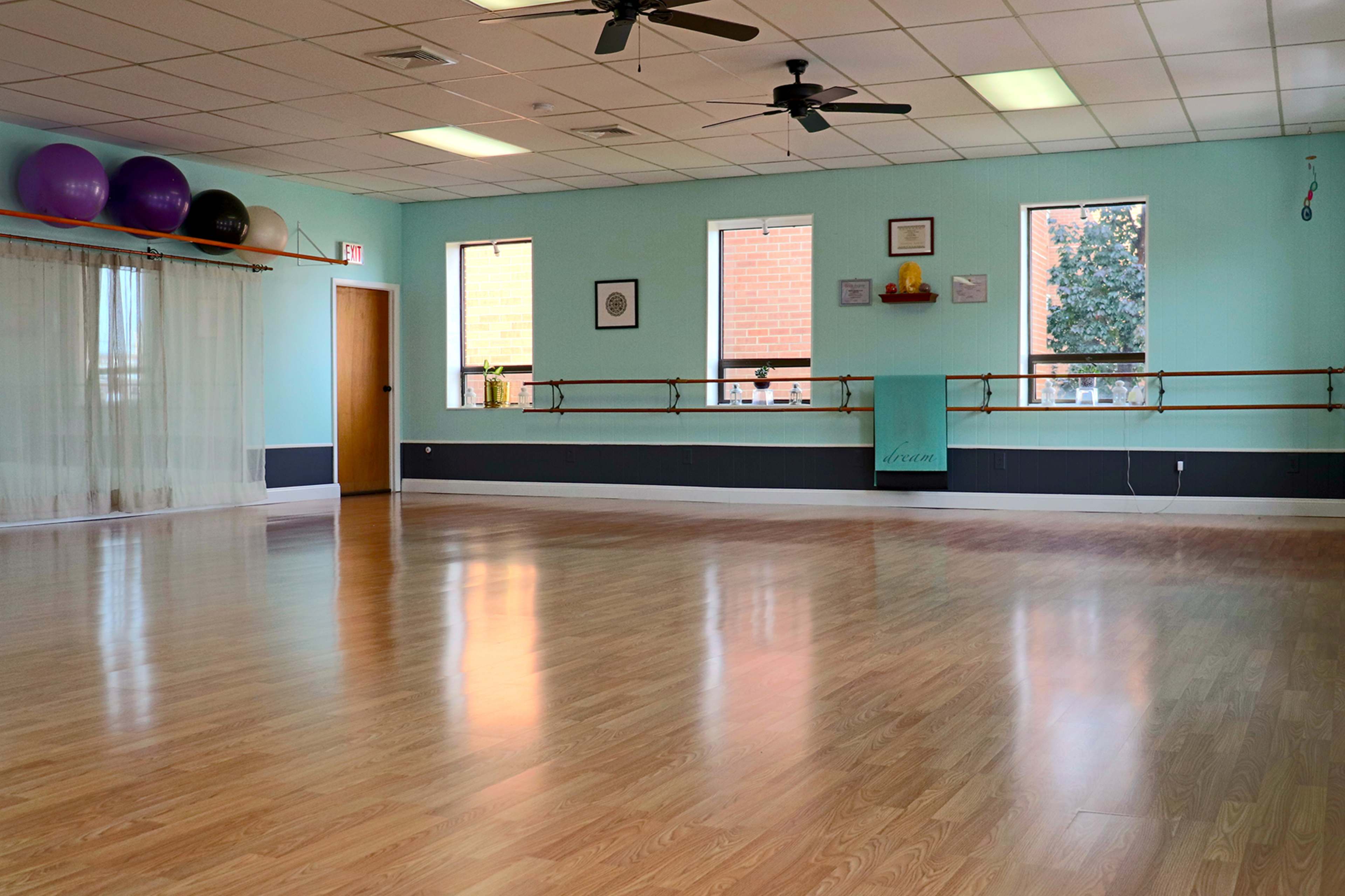 A spacious dance studio features a polished wooden floor, large windows, and exercise balls stored on a rack.