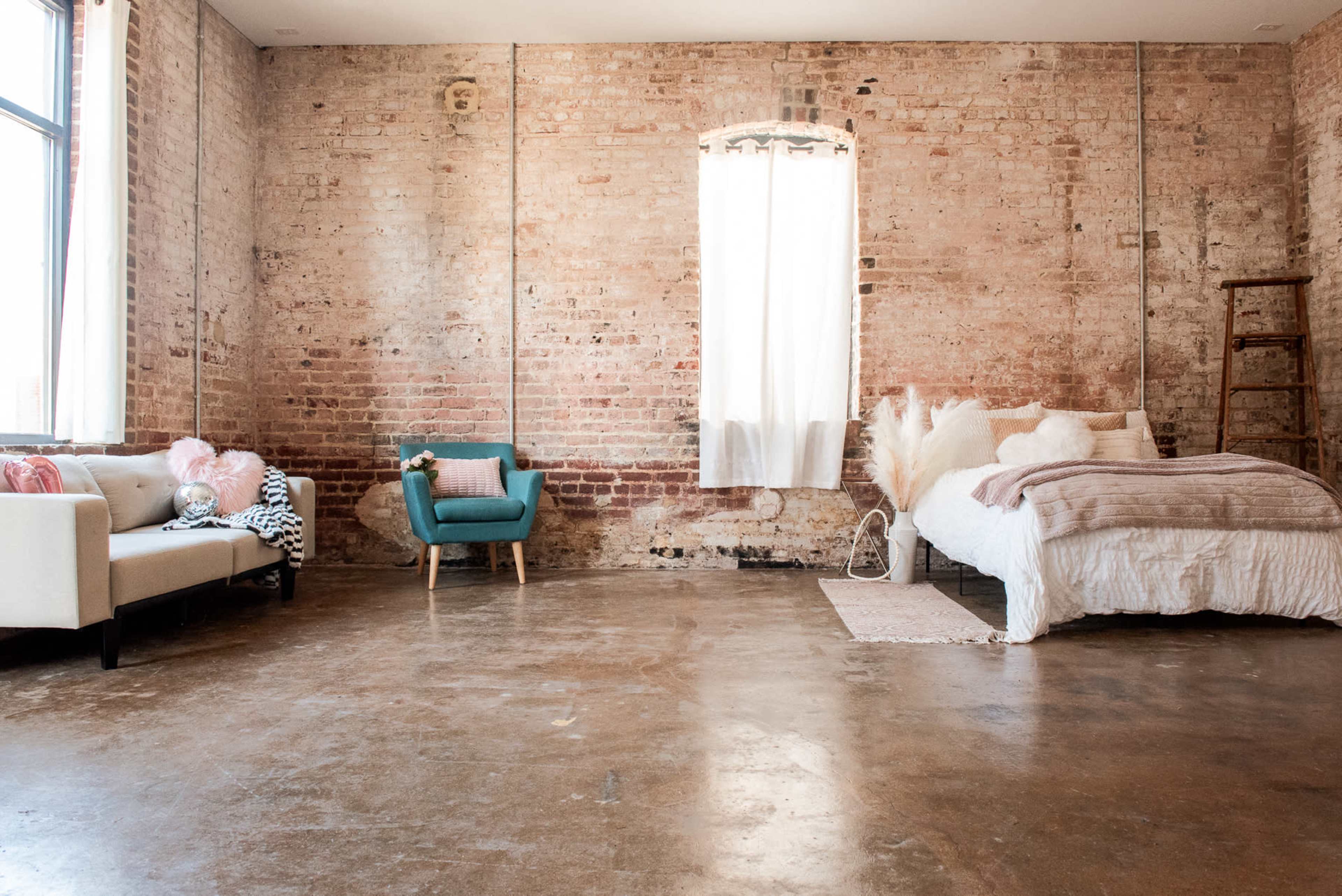 The image shows a spacious room with exposed brick walls, a bed covered in light bedding, a blue accent chair, and a cream-colored sofa.