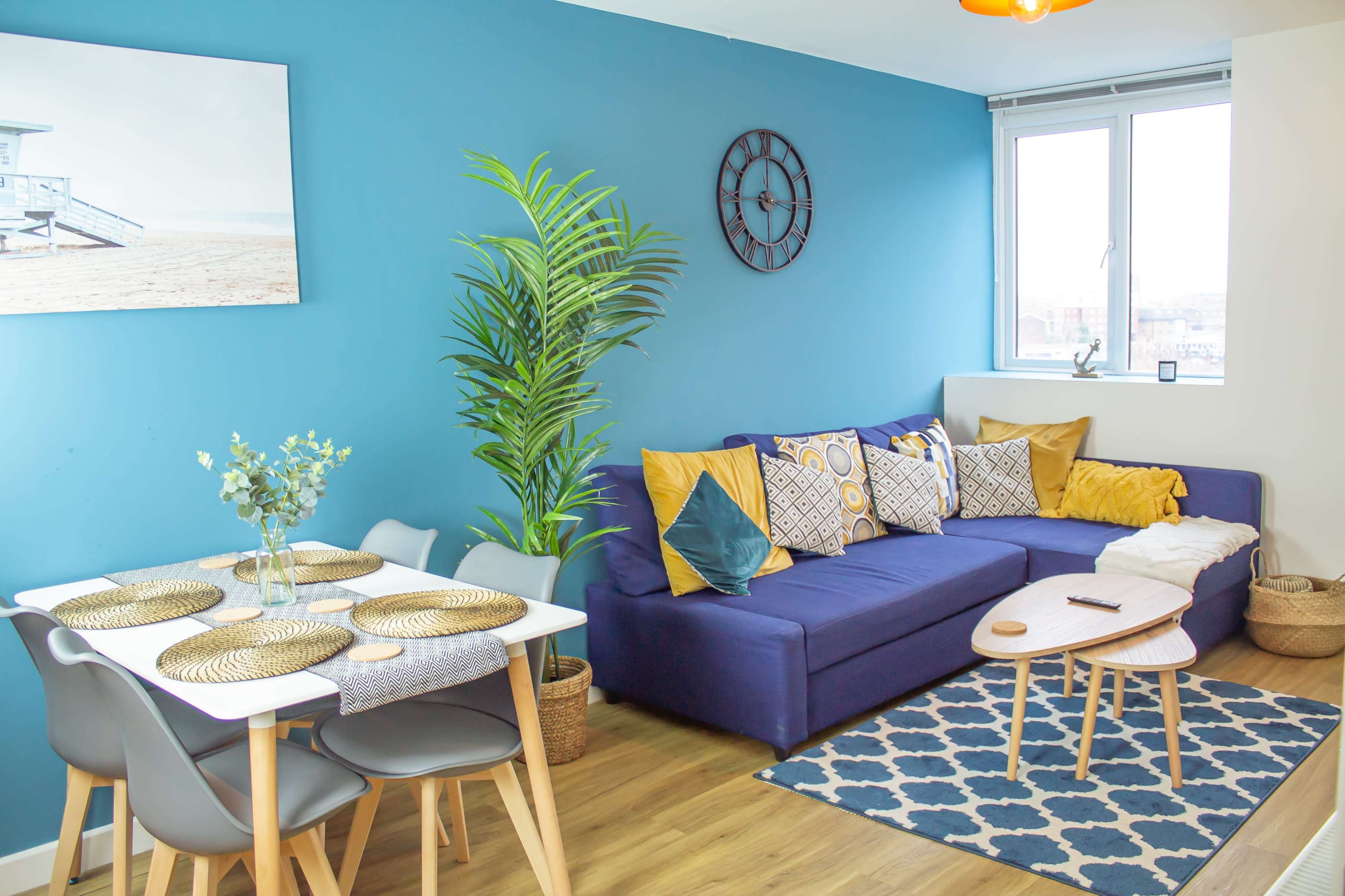 A bright living room features a blue wall, a couch with decorative pillows, a dining table set for four, and a large window allowing natural light.