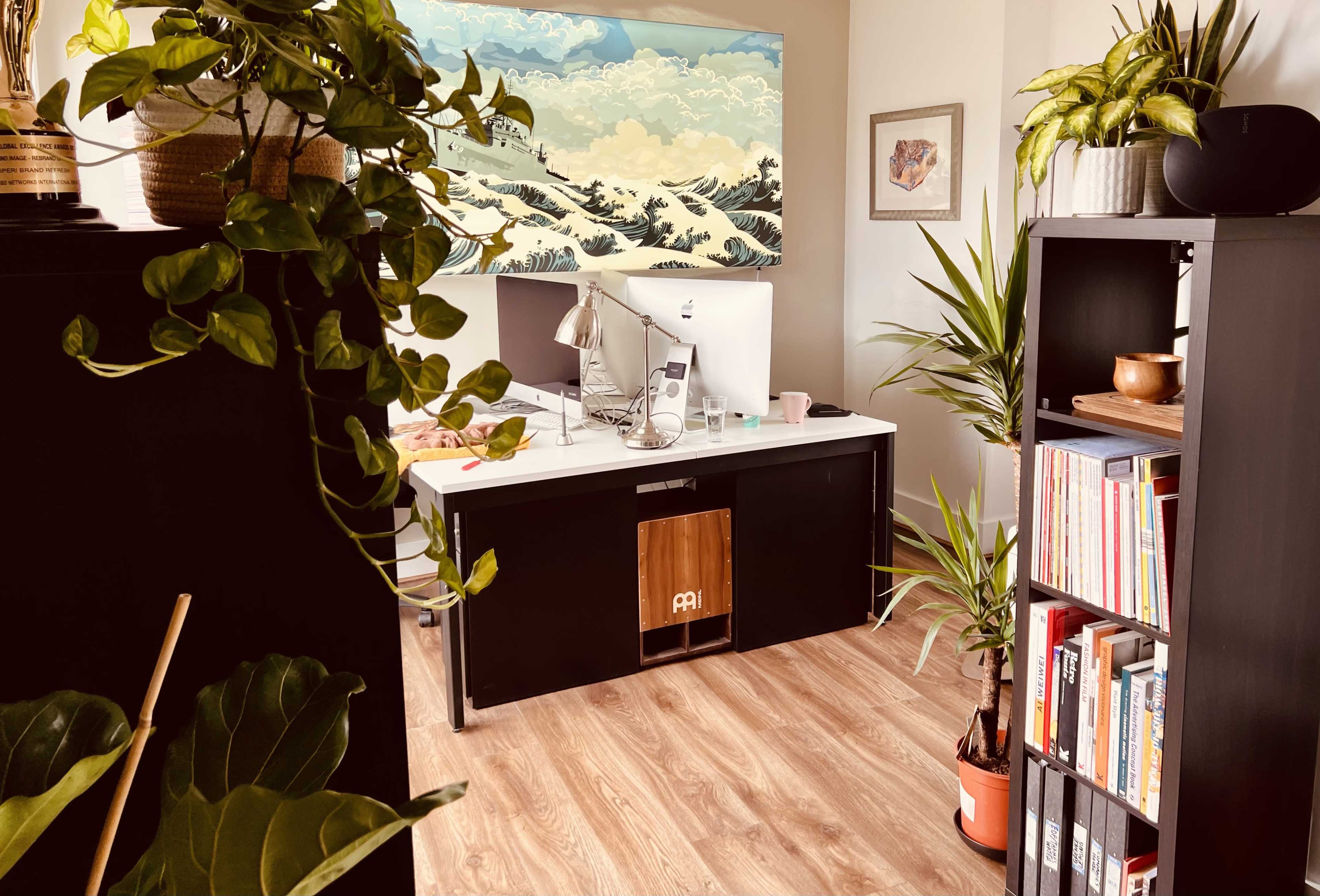 A cozy home office features a desk with a computer and plants, surrounded by bookshelves and artwork on the walls.