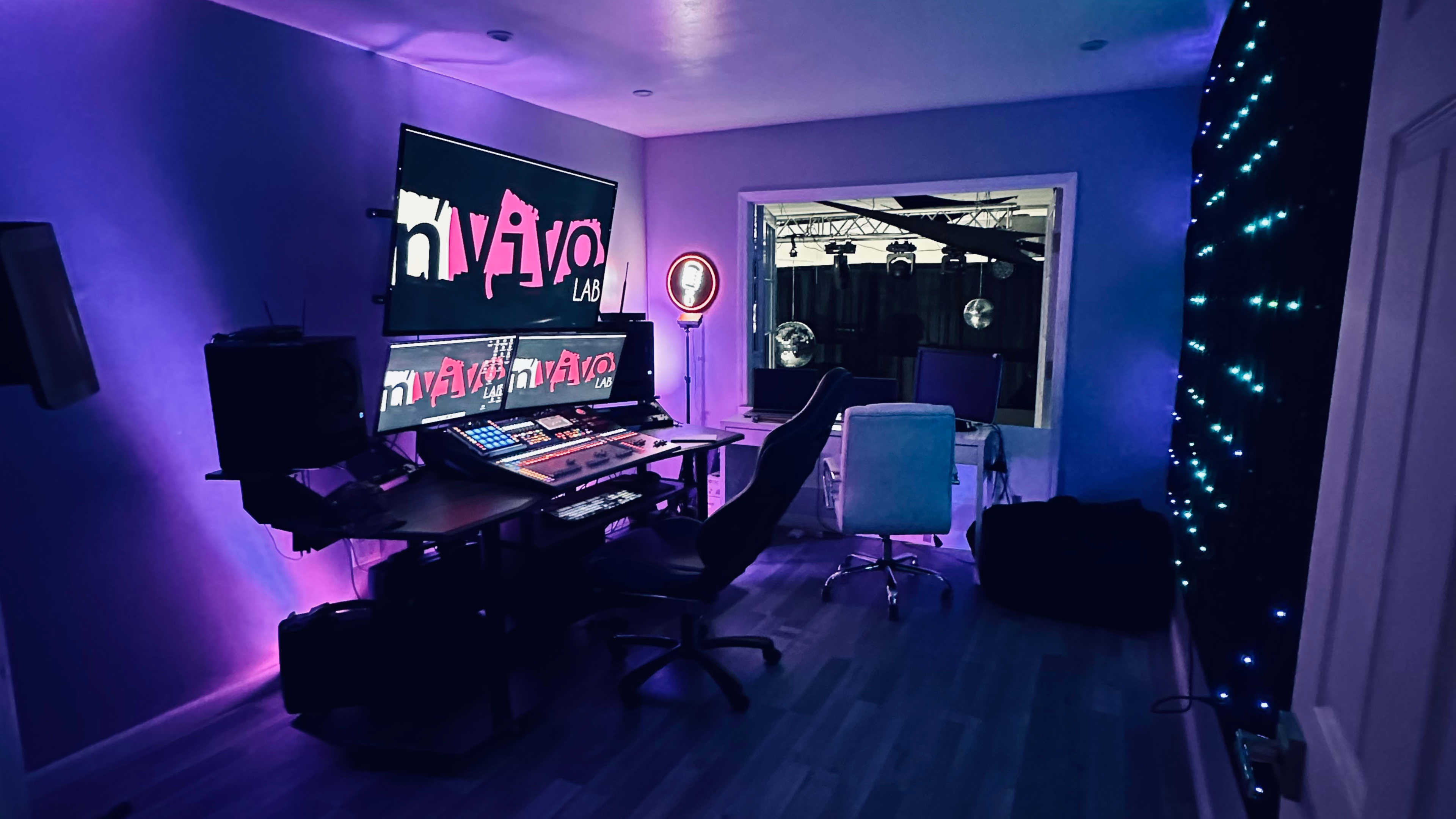 A modern music studio with a control desk featuring multiple screens, chairs, and ambient purple lighting.