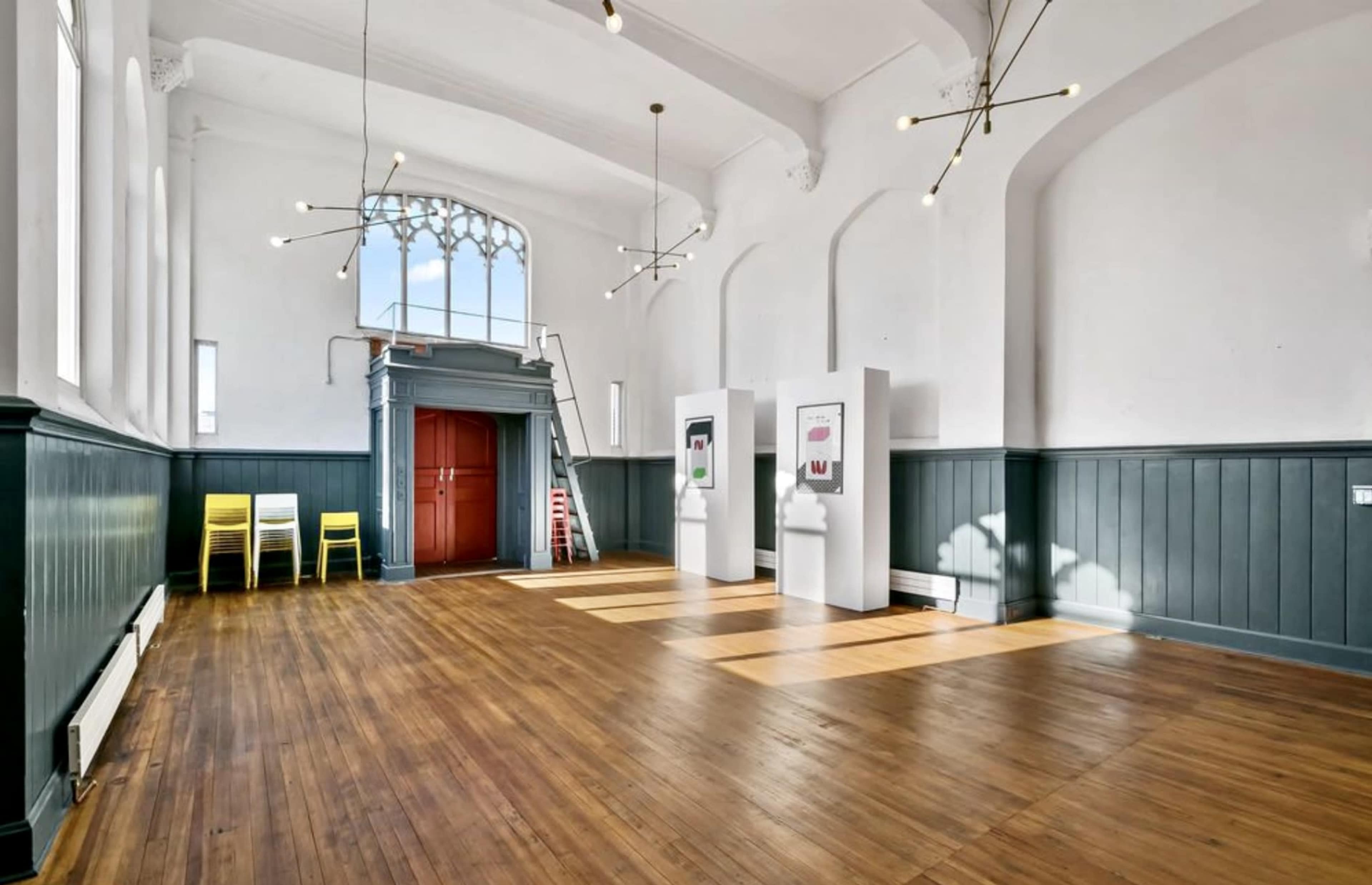 An empty room with wooden floors, bright lighting, and a staircase on one side, adorned with minimalist decor including chairs and artwork.