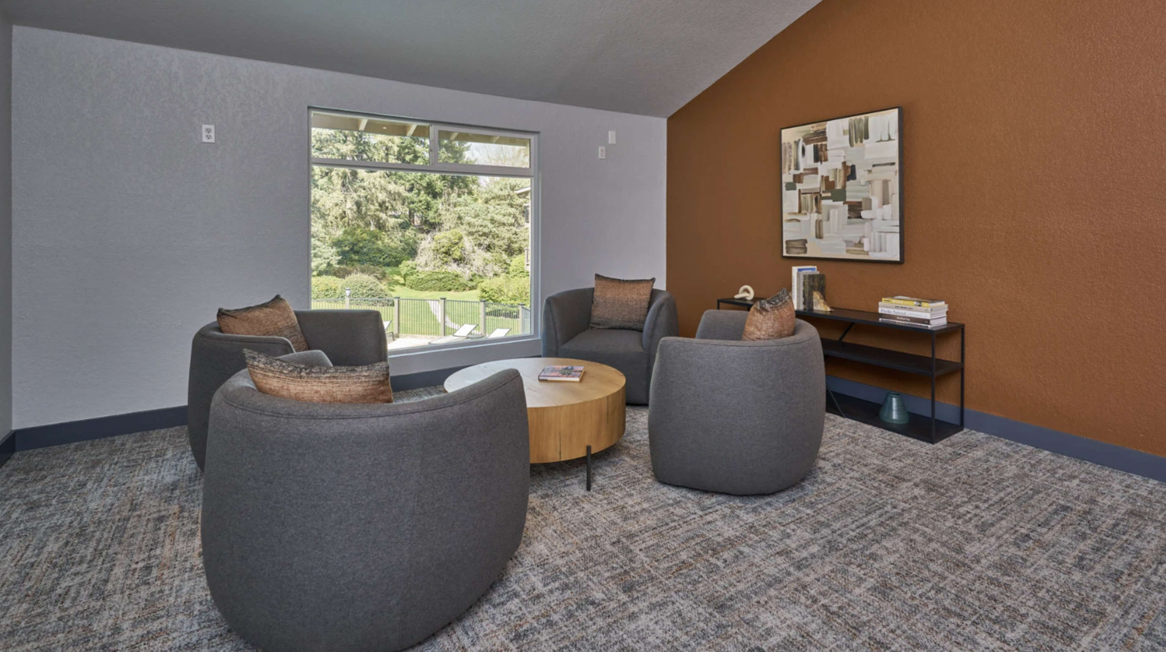 The image shows a cozy seating area with four grey armchairs arranged around a round wooden coffee table, and a large window providing a view of an outdoor landscape.