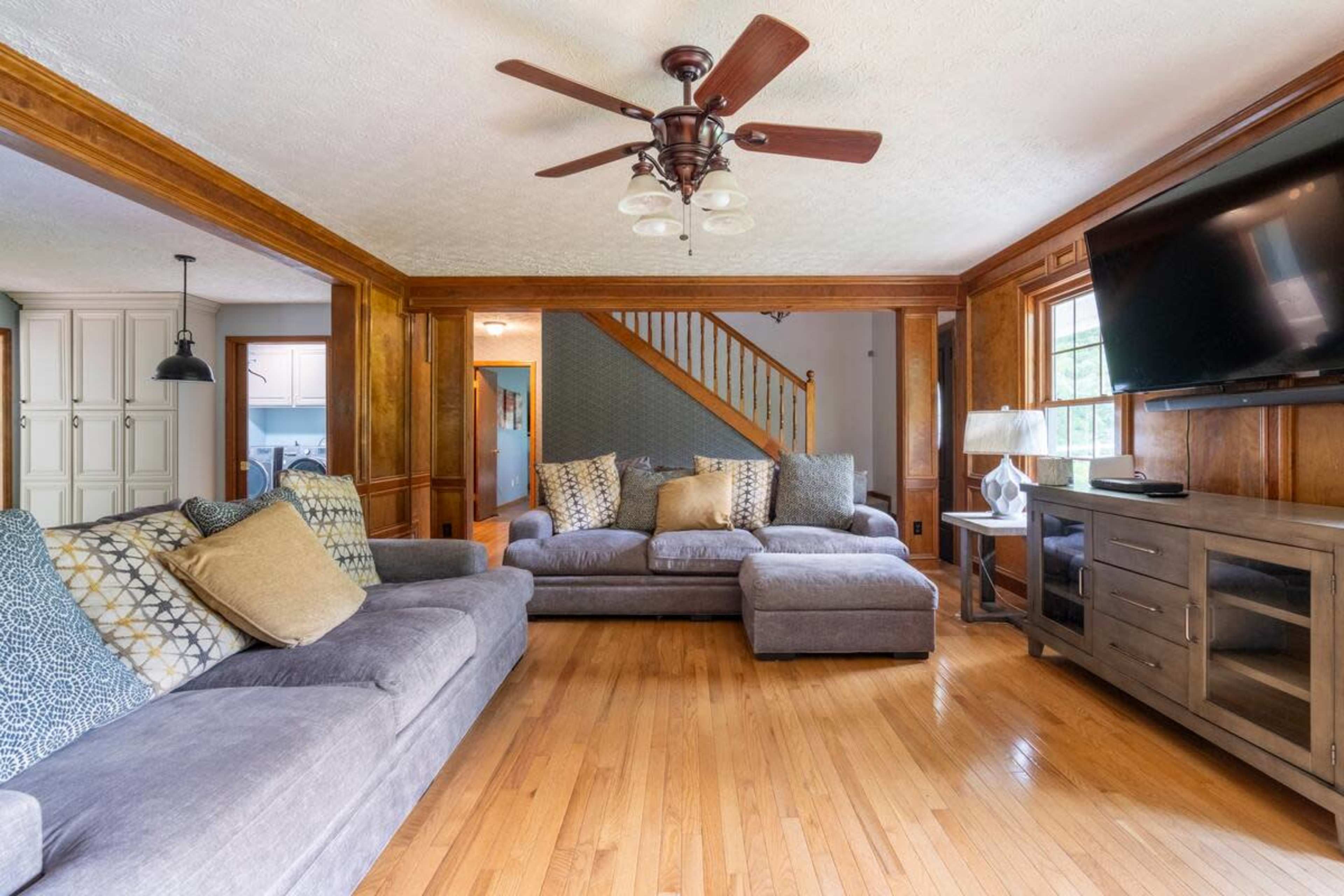A living room features a sectional sofa, wooden accents, a ceiling fan, and a television mounted on a side cabinet.