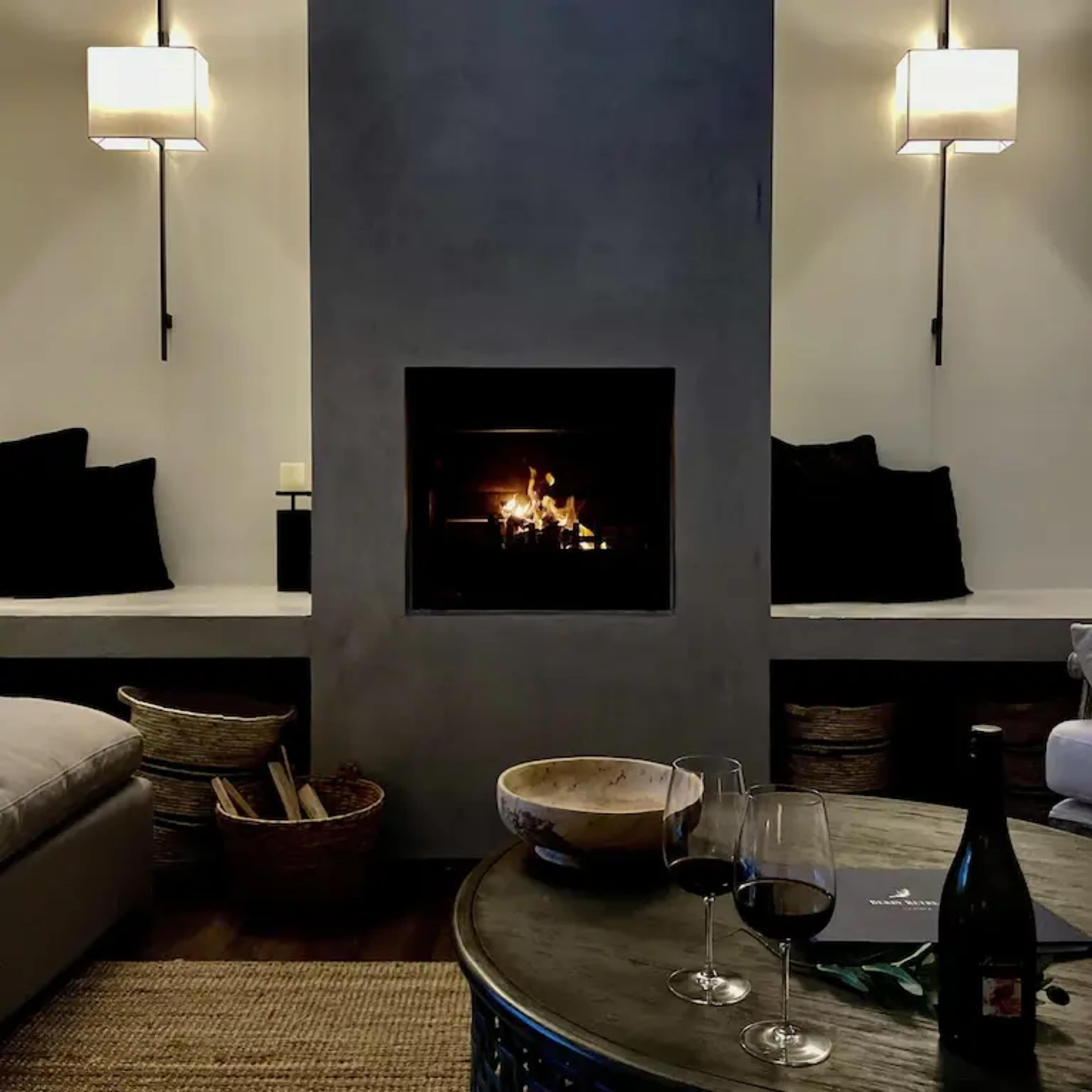 A cozy living room with a fireplace, two wall sconces, a round table holding two glasses of red wine, and a bowl on top, surrounded by dark cushions and a woven rug.