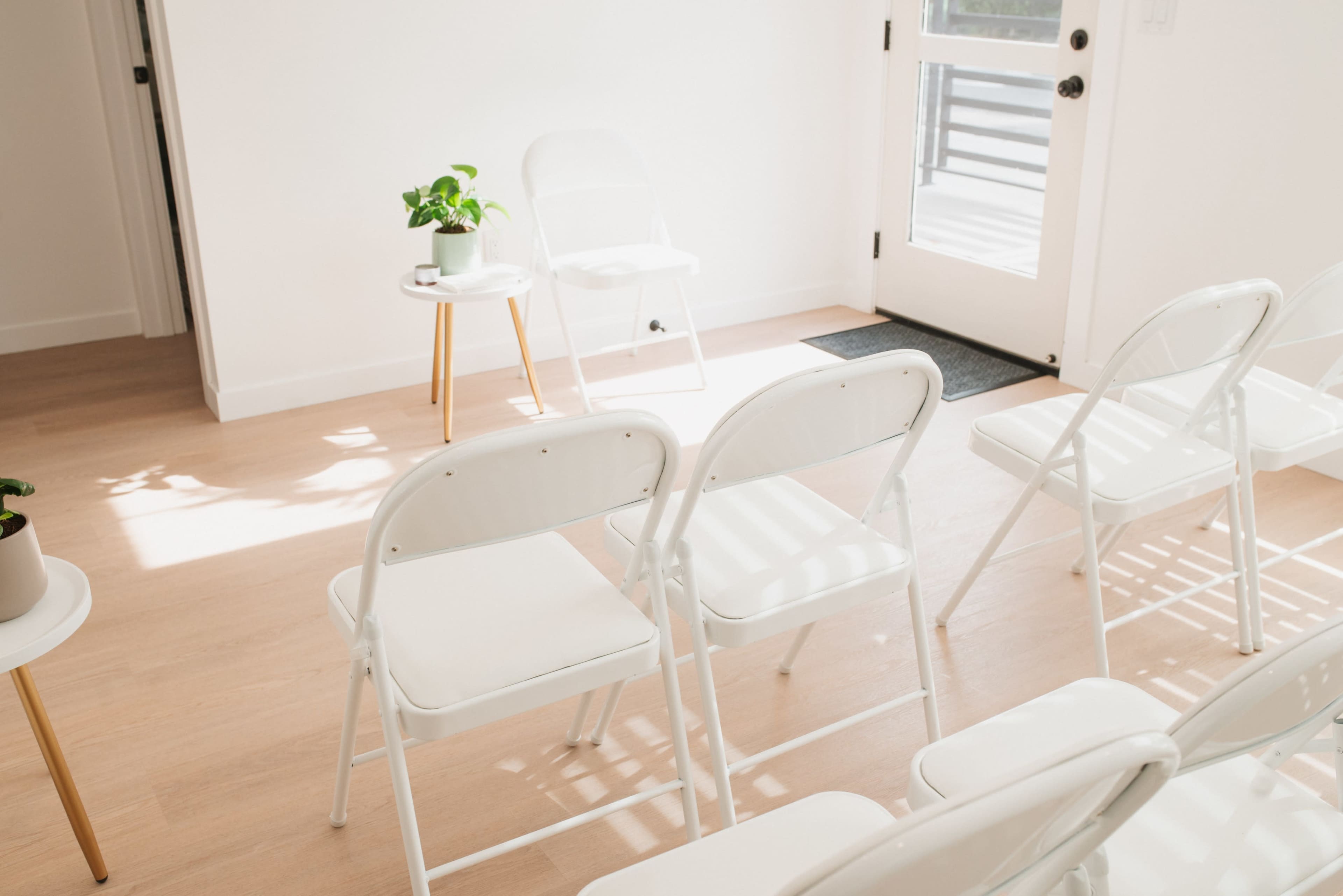 The scene features a bright room with white folding chairs arranged in rows, a small side table with a plant, and a door leading outside.