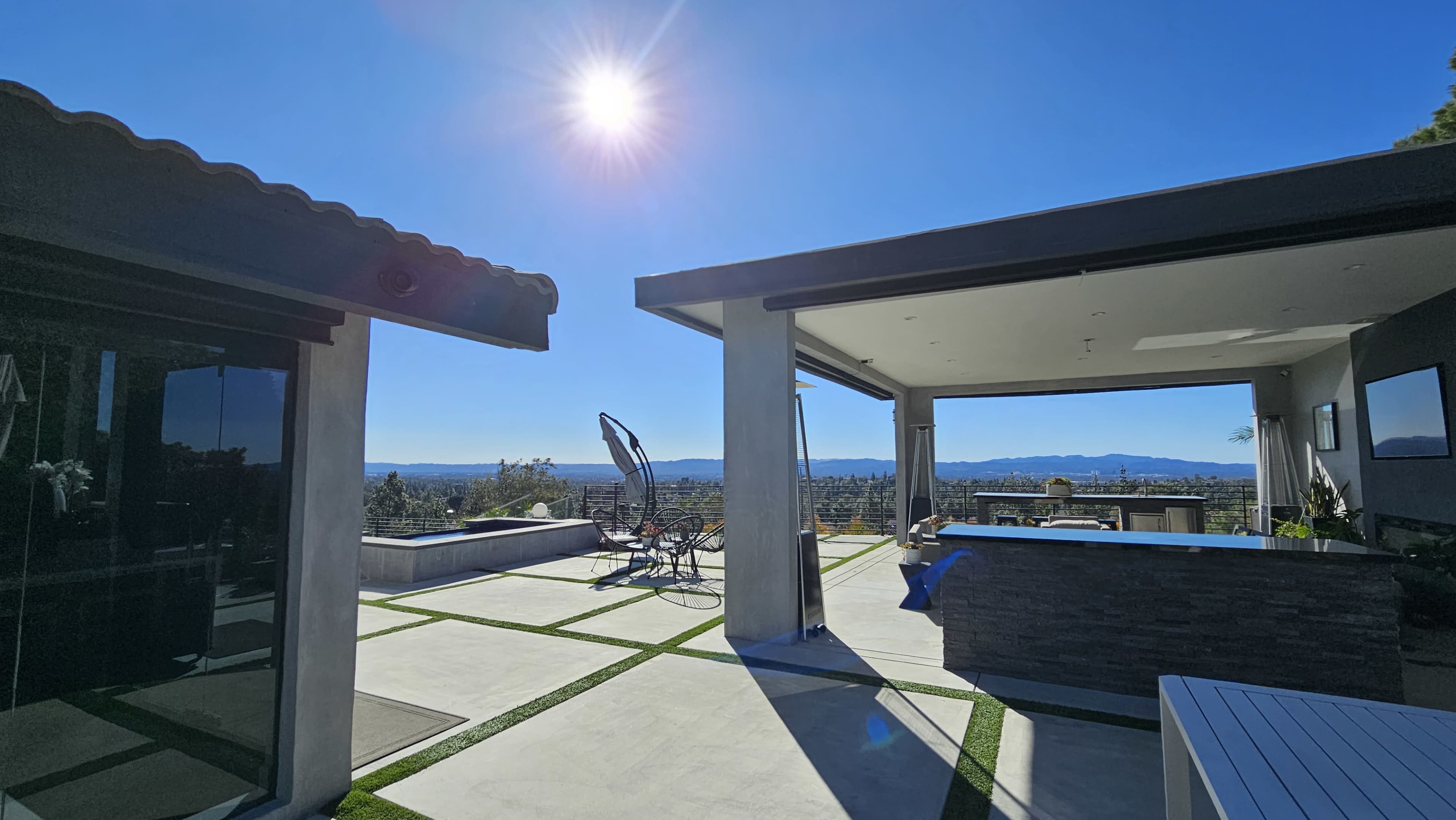 A modern outdoor patio overlooks a panoramic view of a valley under a clear blue sky, with a sun shining brightly.