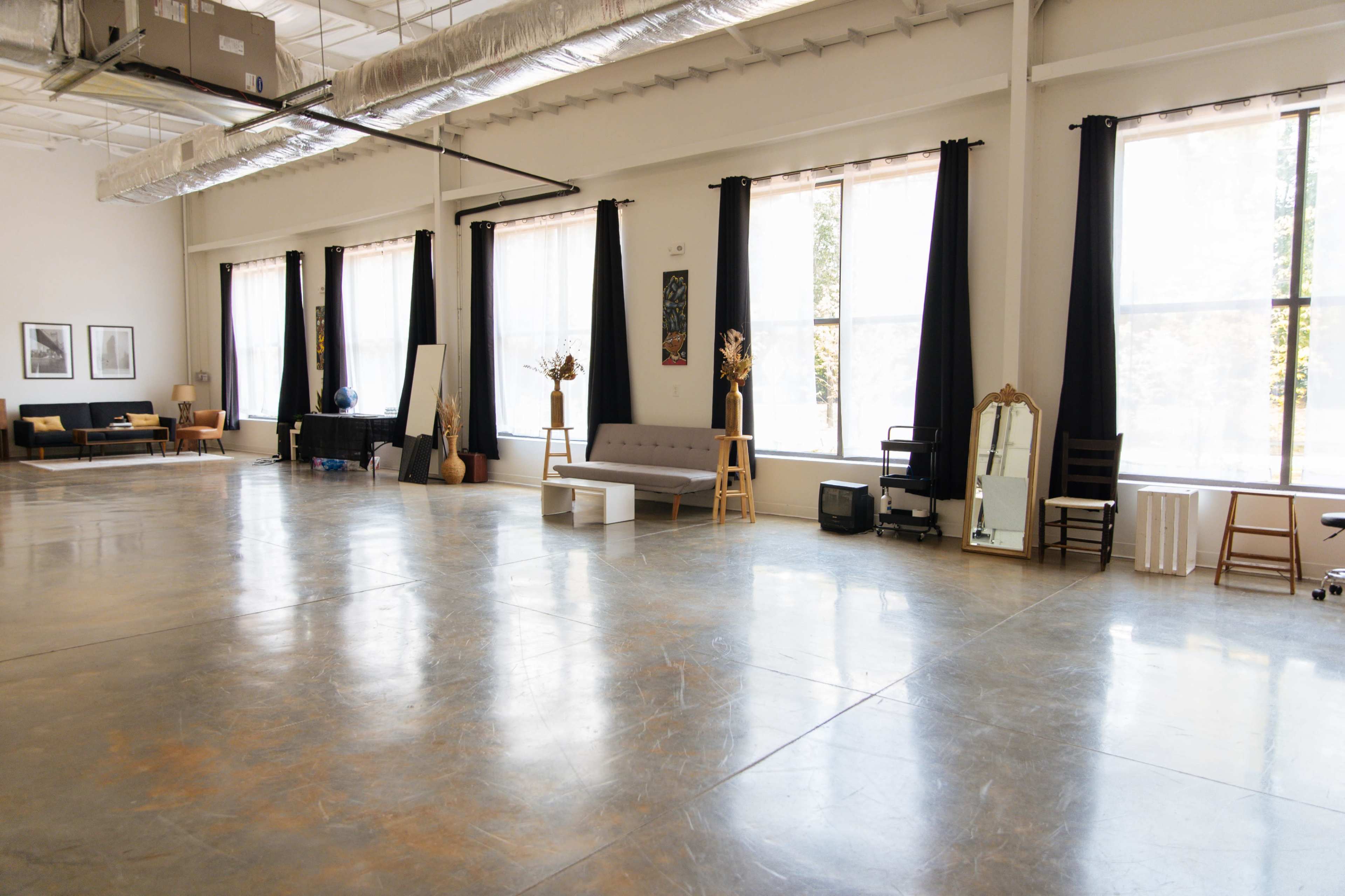 A spacious, well-lit room with large windows, featuring minimal furniture including a gray sofa, a mirror, and decorative elements.