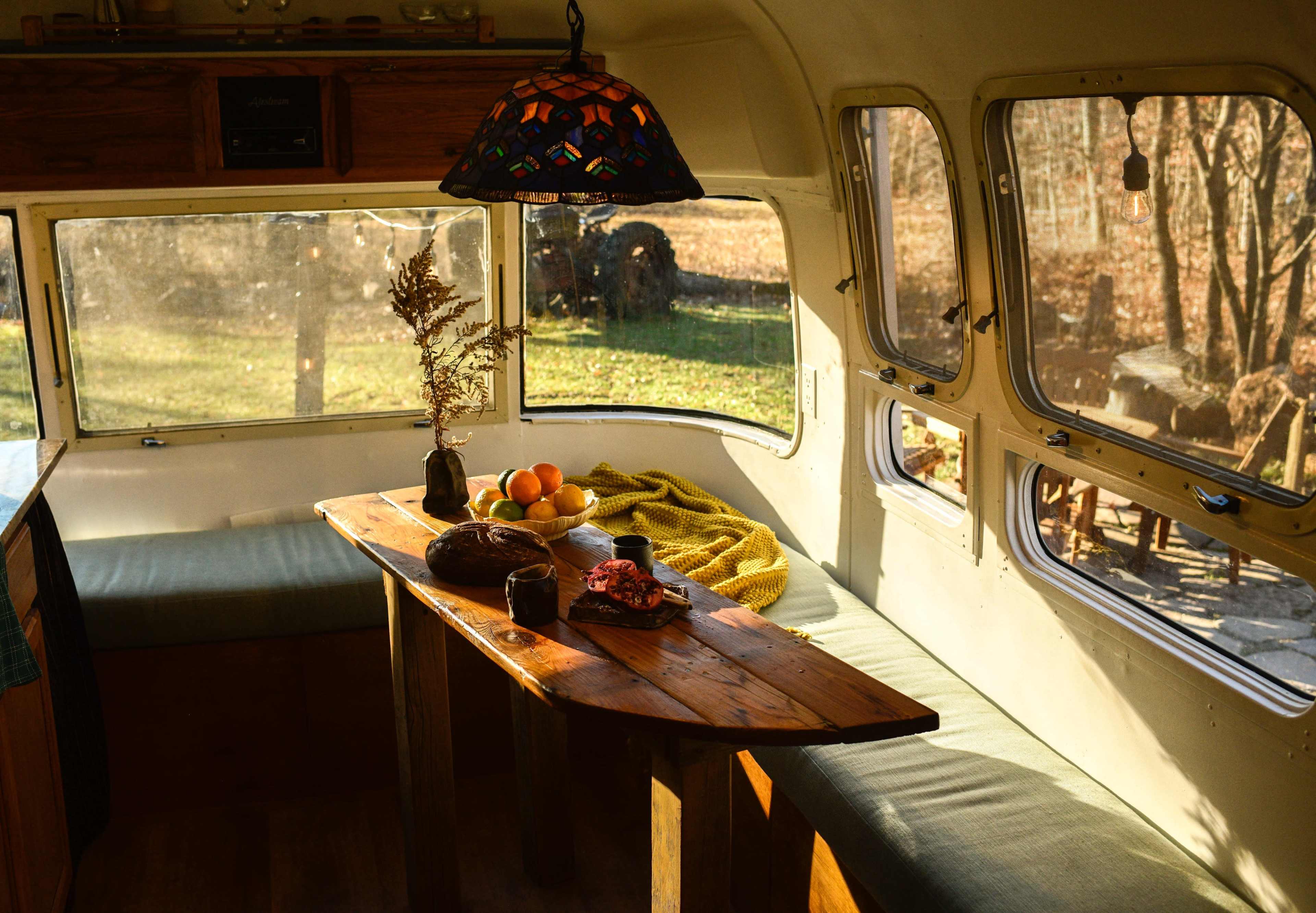 A cozy interior of a camper features a wooden table with fruit and a loaf of bread, surrounded by natural light from large windows.