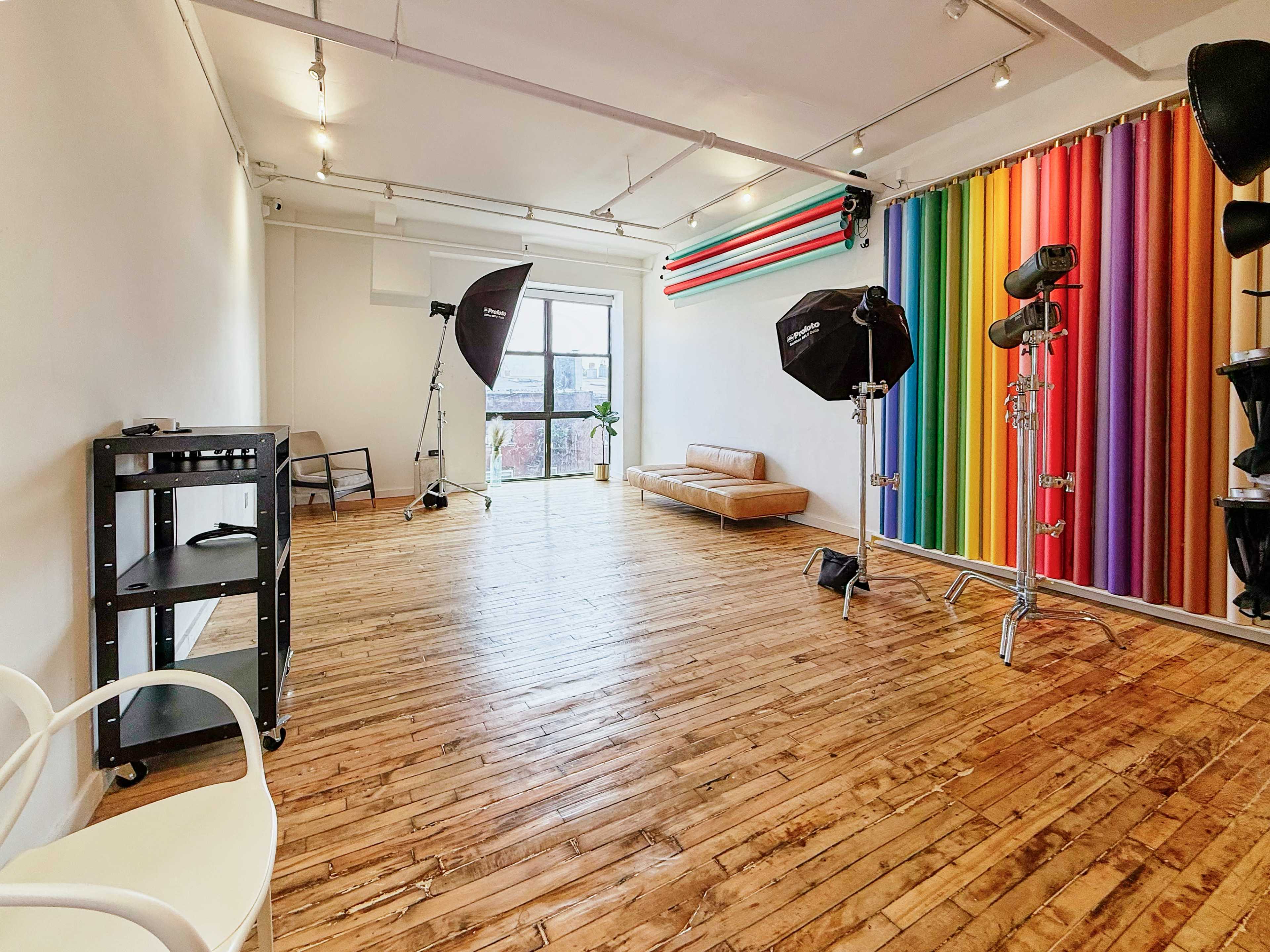 The image shows a spacious photography studio with wooden floors, colorful backdrop curtains, and professional lighting equipment set up.