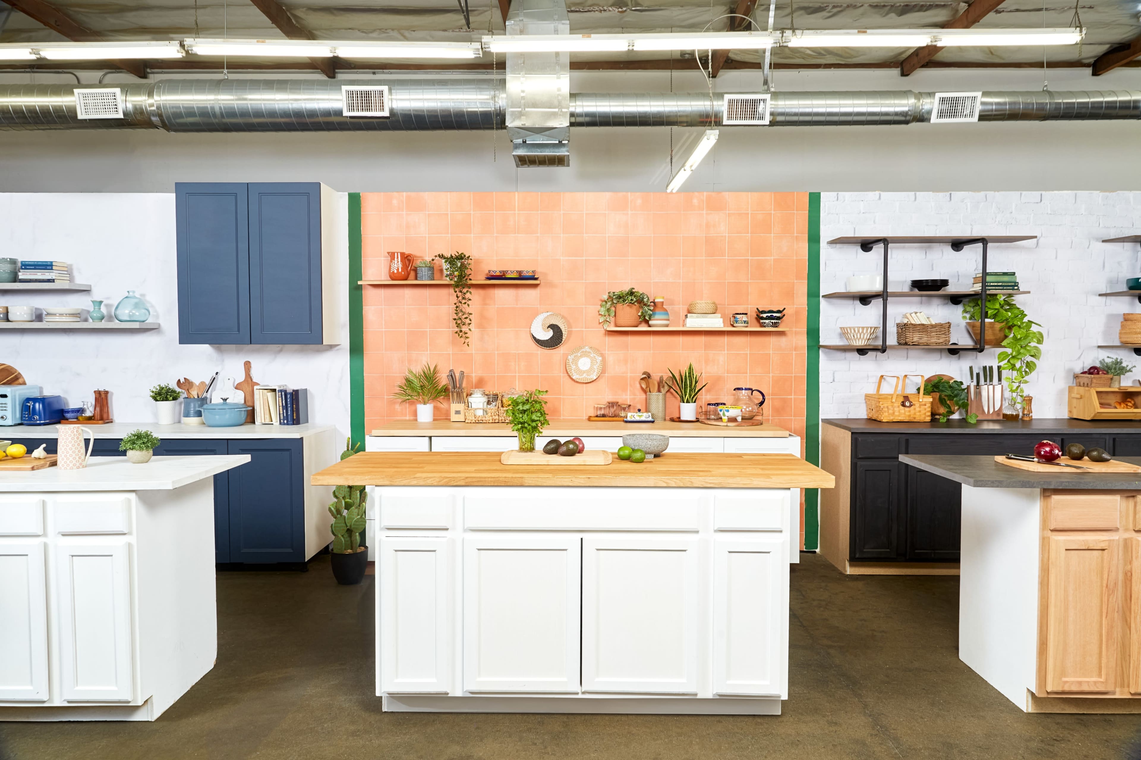A modern kitchen setup featuring multiple islands and cabinets in various colors, with open shelving displaying kitchenware and plants against a tiled backdrop.