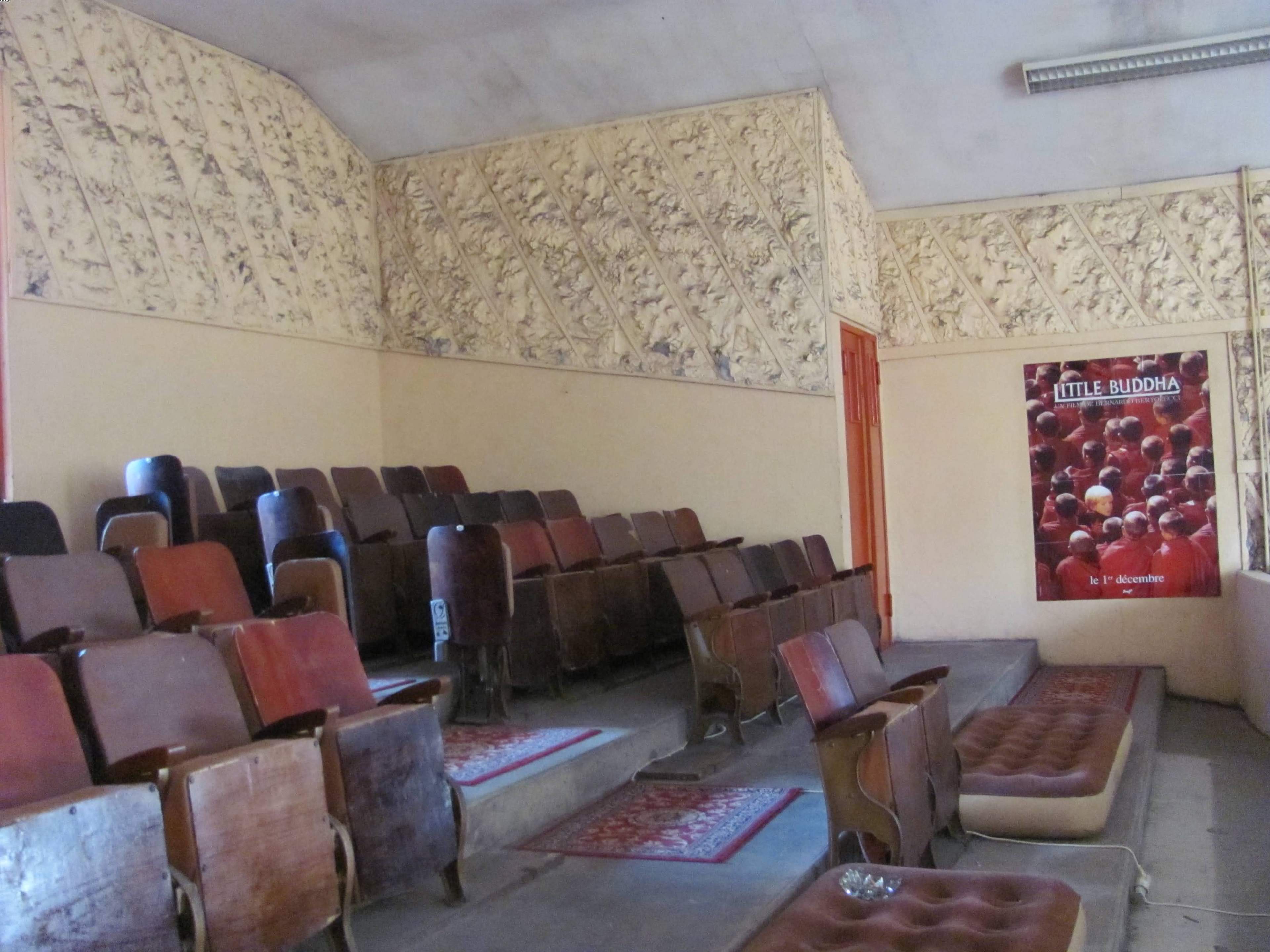 A sparsely furnished room with wooden seating, decorative wall patterns, and a poster titled "Little Buddha" displayed prominently.