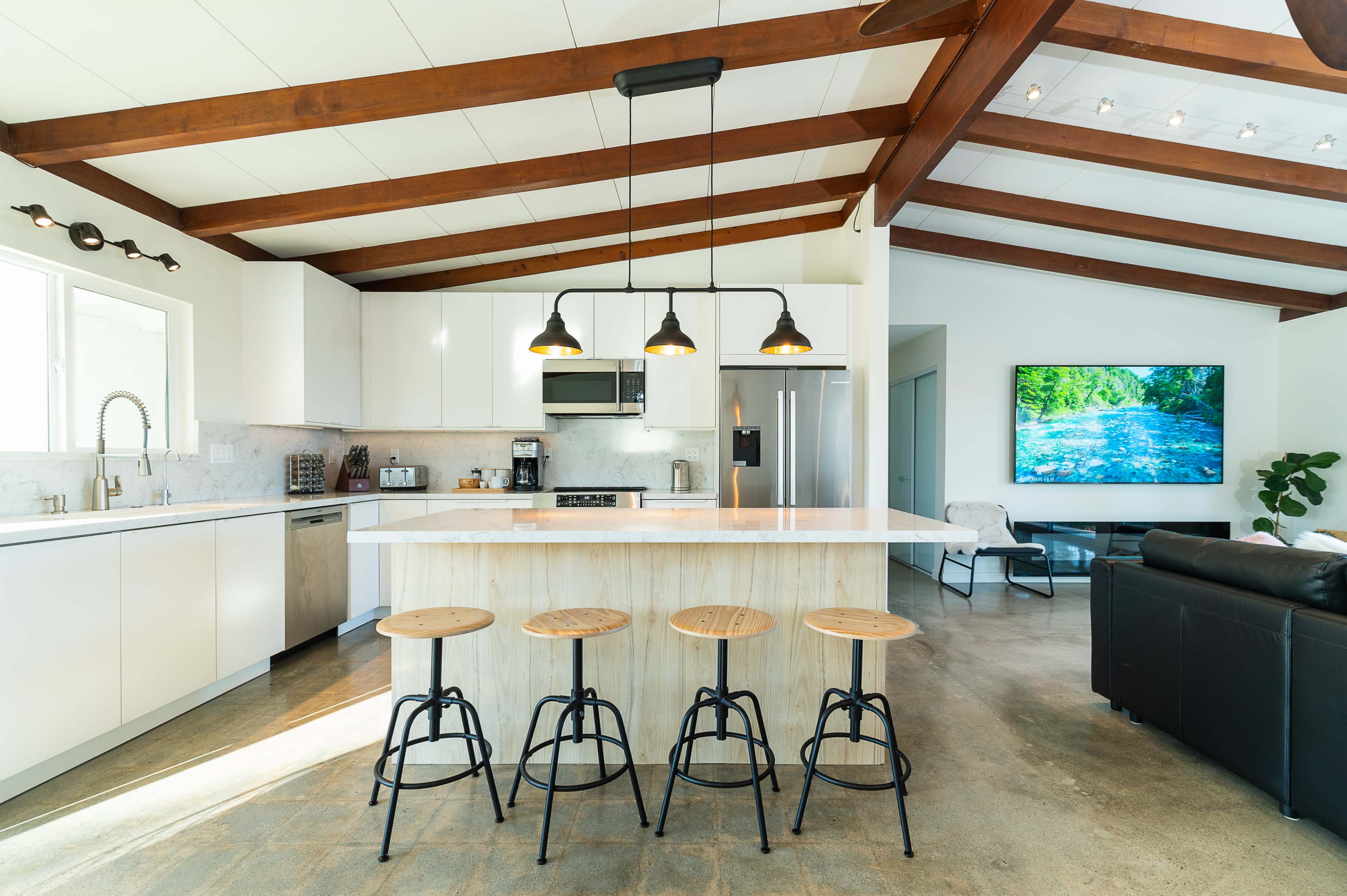 A modern kitchen features a large island with three wooden stools, white cabinetry, stainless steel appliances, and exposed wooden beams above.