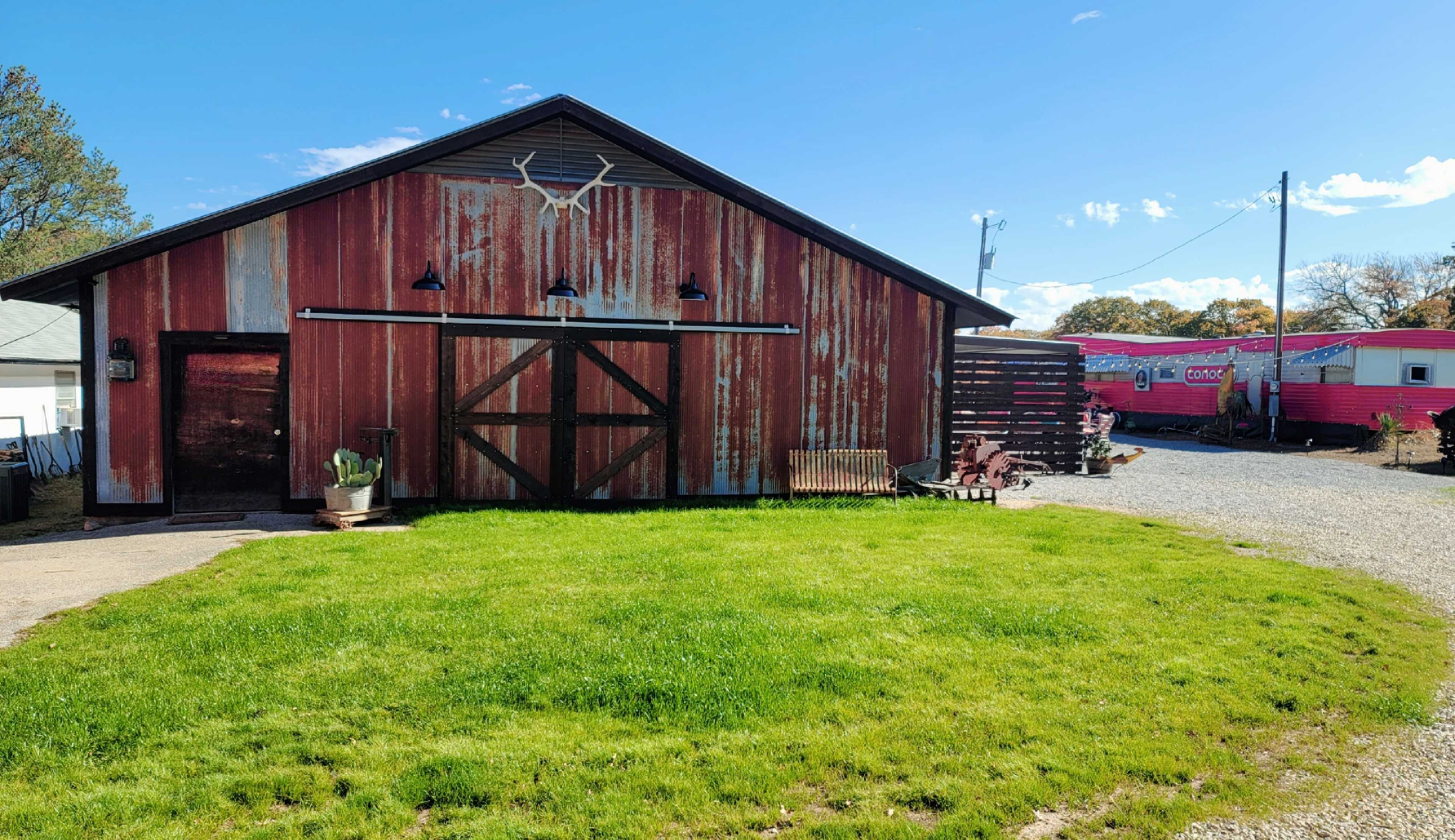 A weathered red barn with antlers on the facade stands next to a pink trailer in a grassy area.