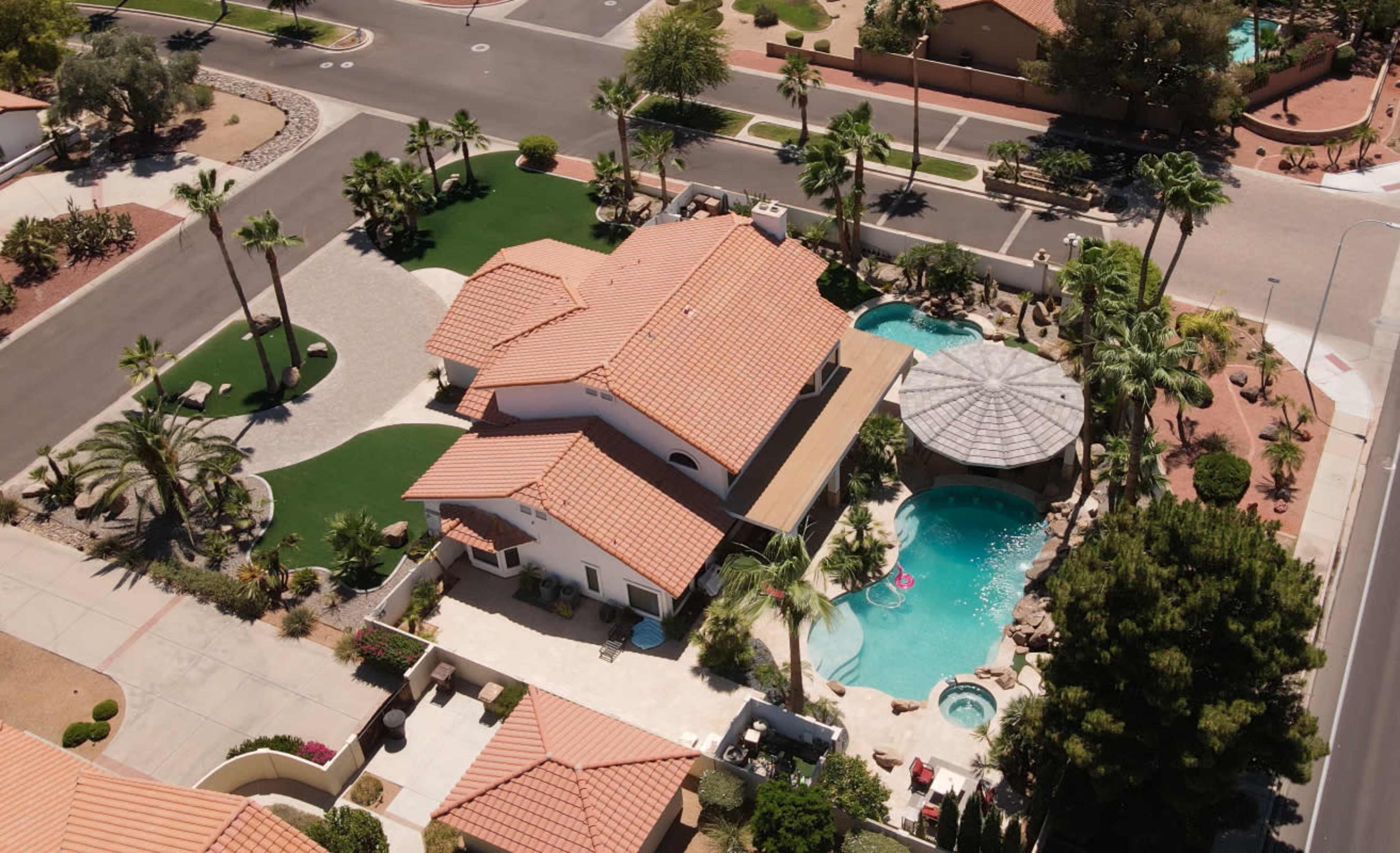 An aerial view of a residential property featuring a house with a tiled roof, a swimming pool, landscaped areas, and surrounding palm trees.