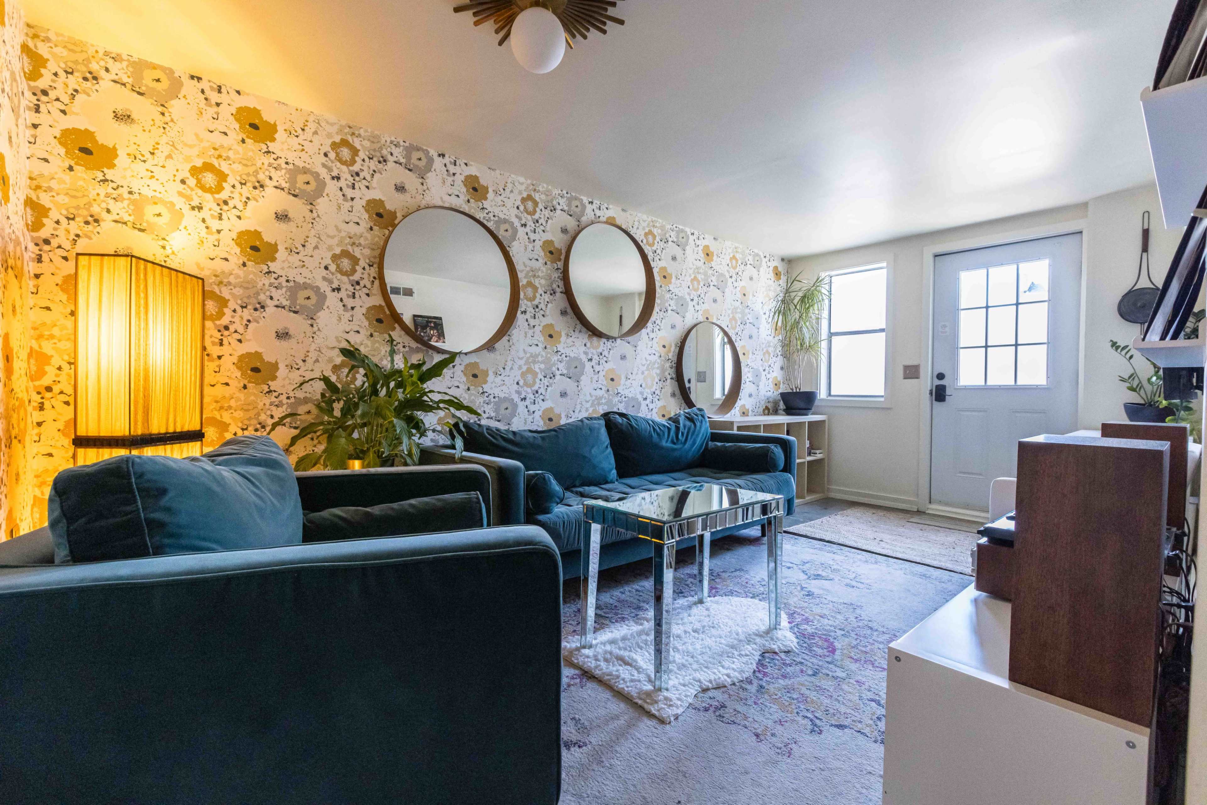 A living room features teal sofas, a glass coffee table, three round mirrors on the floral wallpaper, and a doorway with a window.