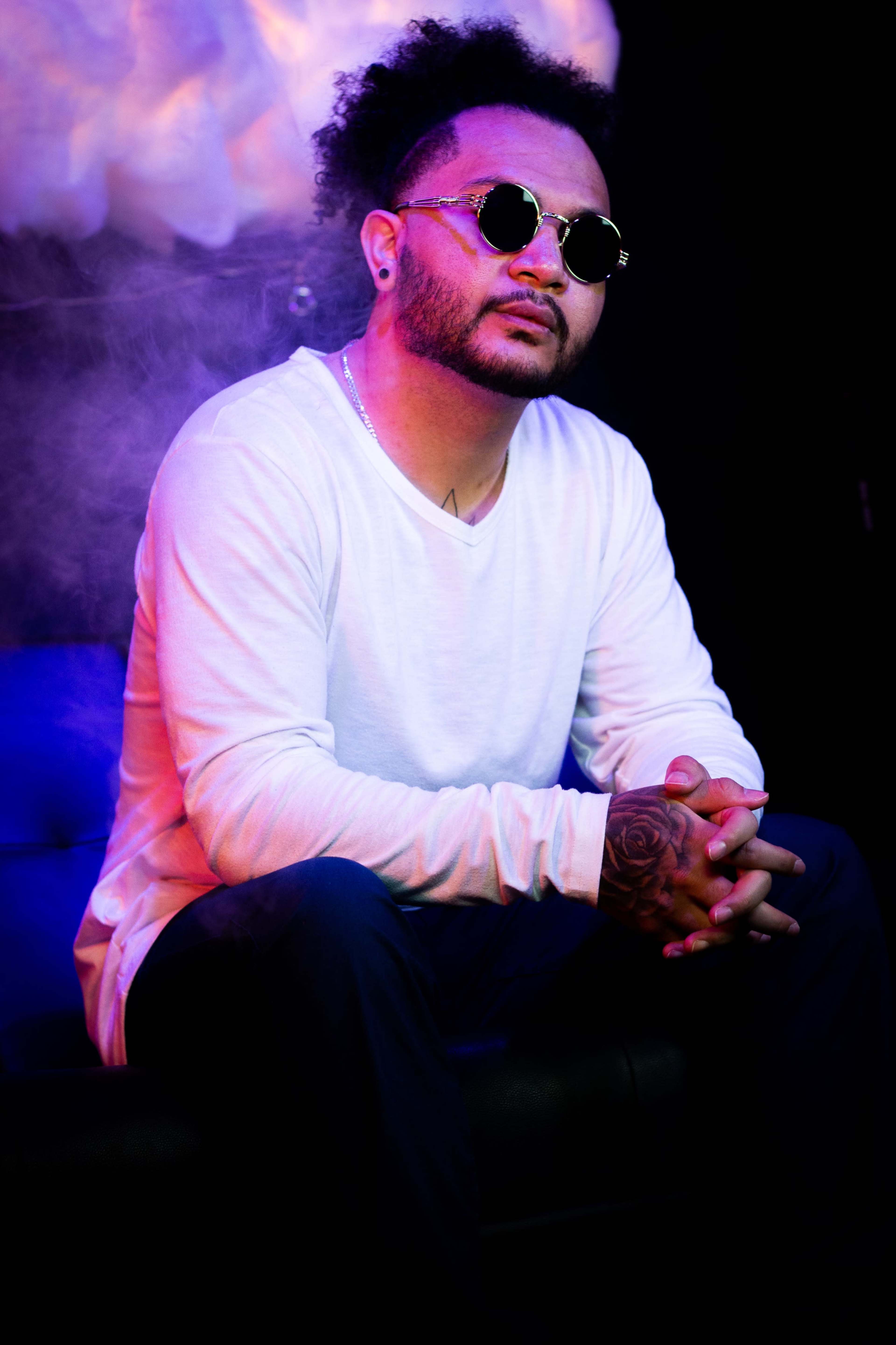 A man with curly hair, wearing sunglasses and a white long-sleeve shirt, sits on a couch with his hands clasped in front of him amid a misty, colorful background.