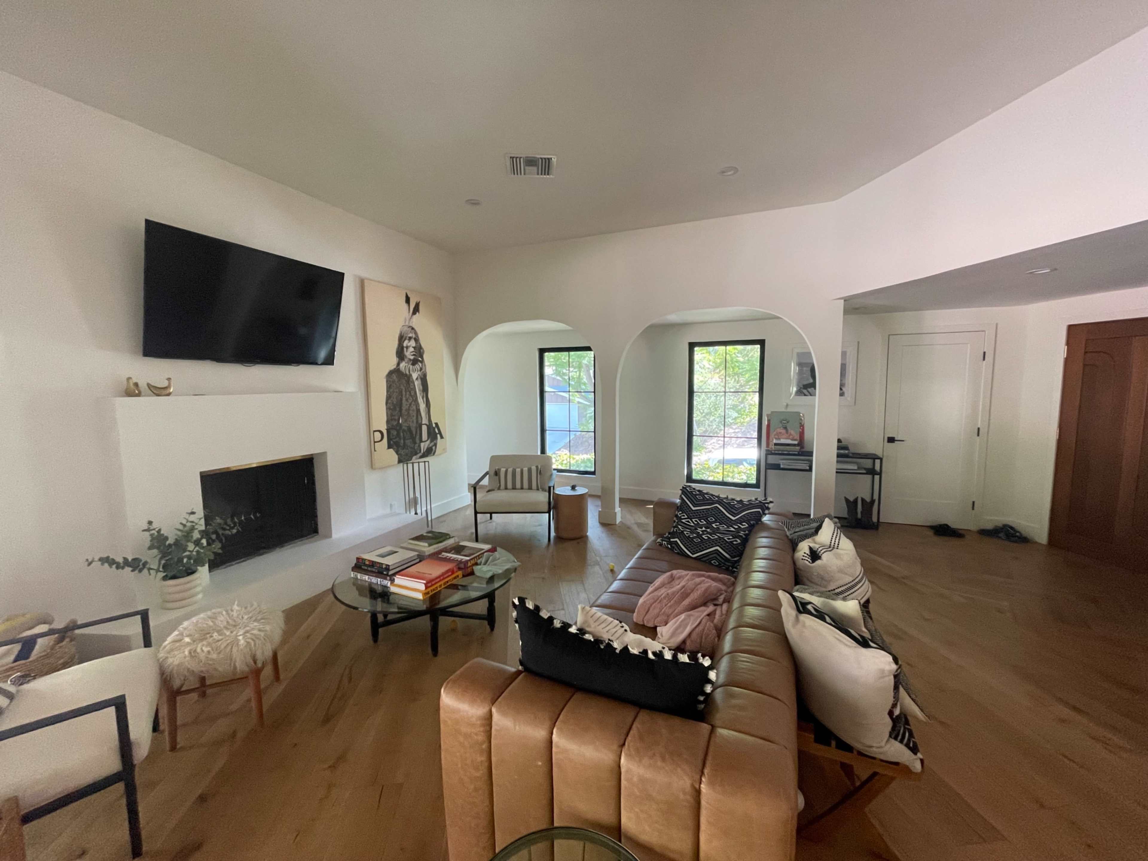 A living room features a leather sofa, coffee table with books, and a wall-mounted television, with large windows allowing natural light to enter.