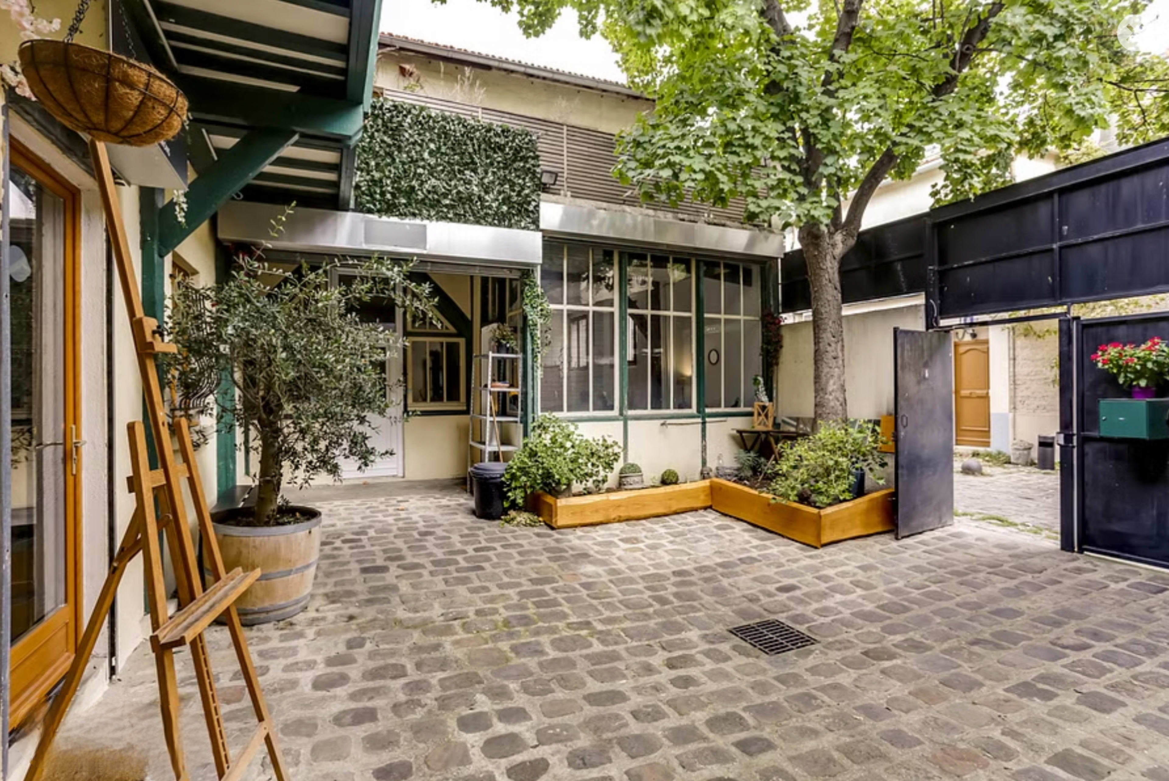 A serene courtyard featuring cobblestone flooring, potted plants, and a large tree, enclosed by buildings with large windows.