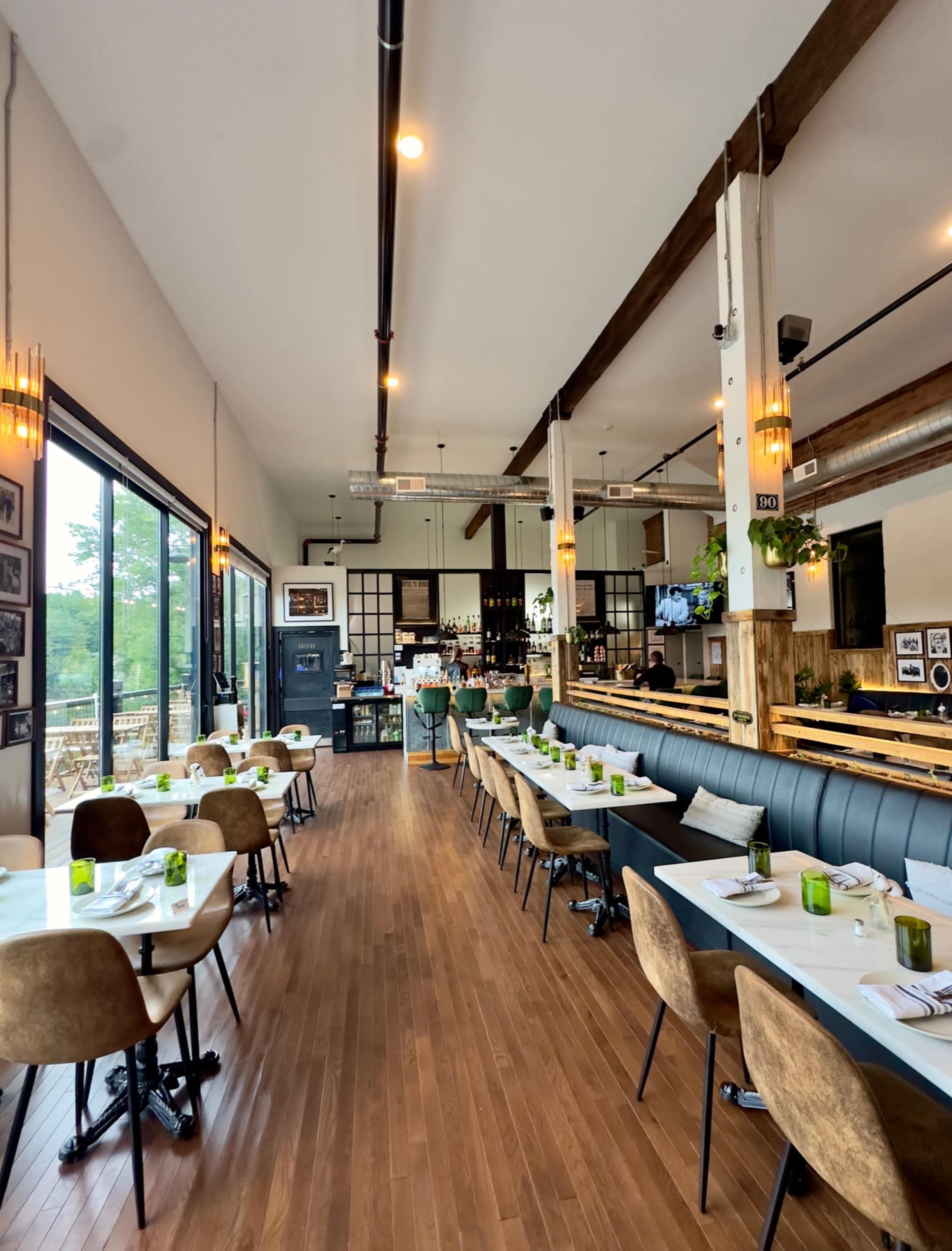 The image shows a modern restaurant interior with wooden floors, elegant seating arrangements, and a bar area in the background.