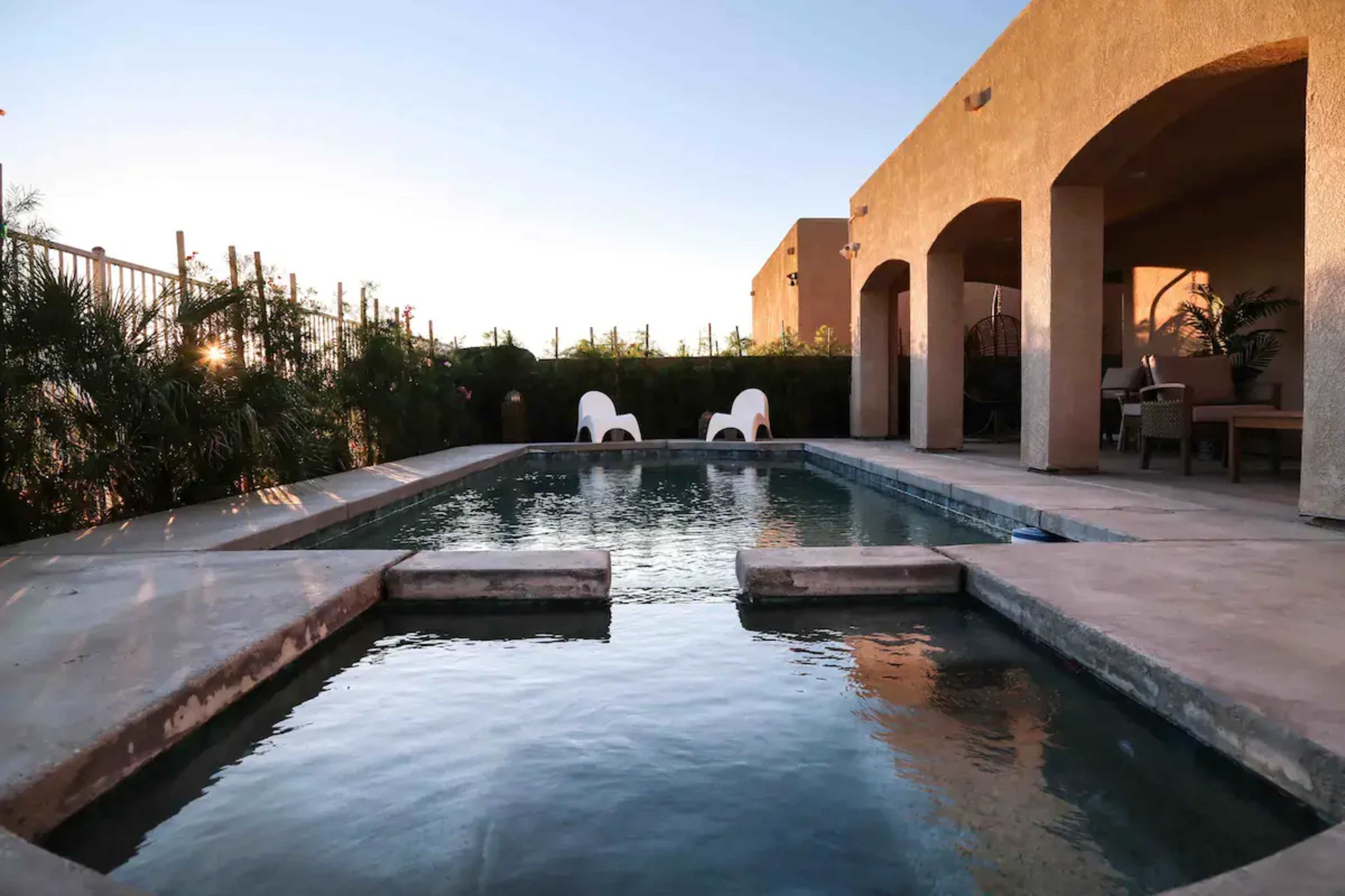 A rectangular swimming pool is surrounded by beige walls and greenery, with two white lounge chairs positioned nearby.