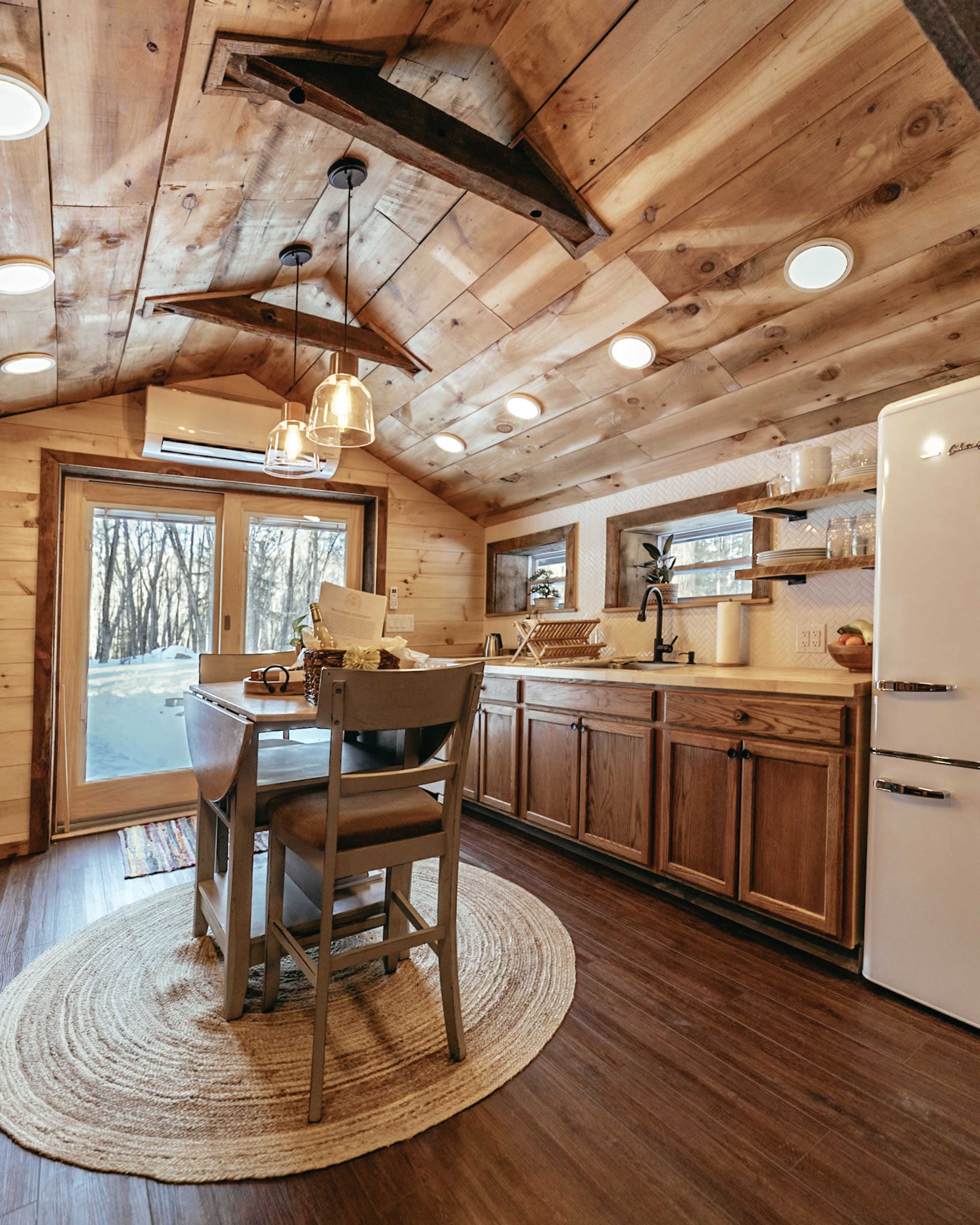 A cozy kitchen with wooden cabinetry, a round dining table, and large windows overlooking a snowy landscape.