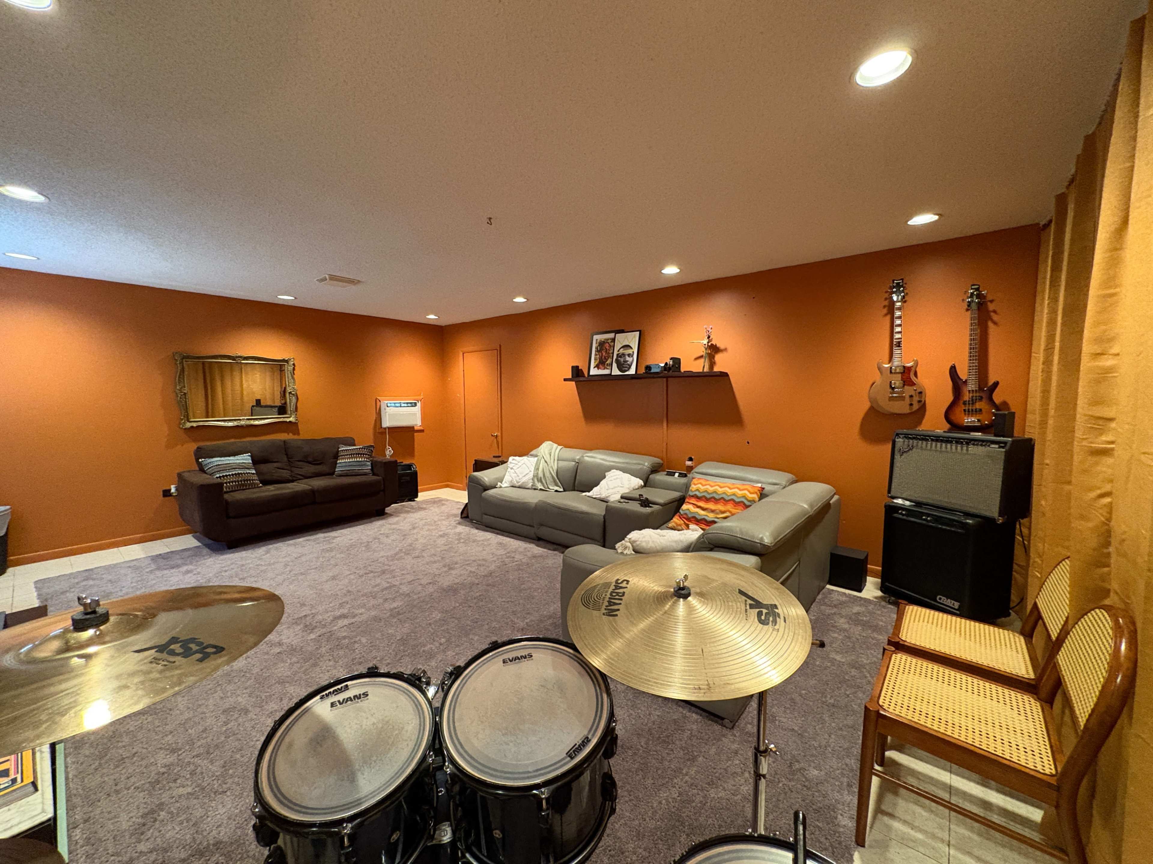 The image shows a finished basement with a seating area, musical instruments on the wall, and a drum set in the foreground.